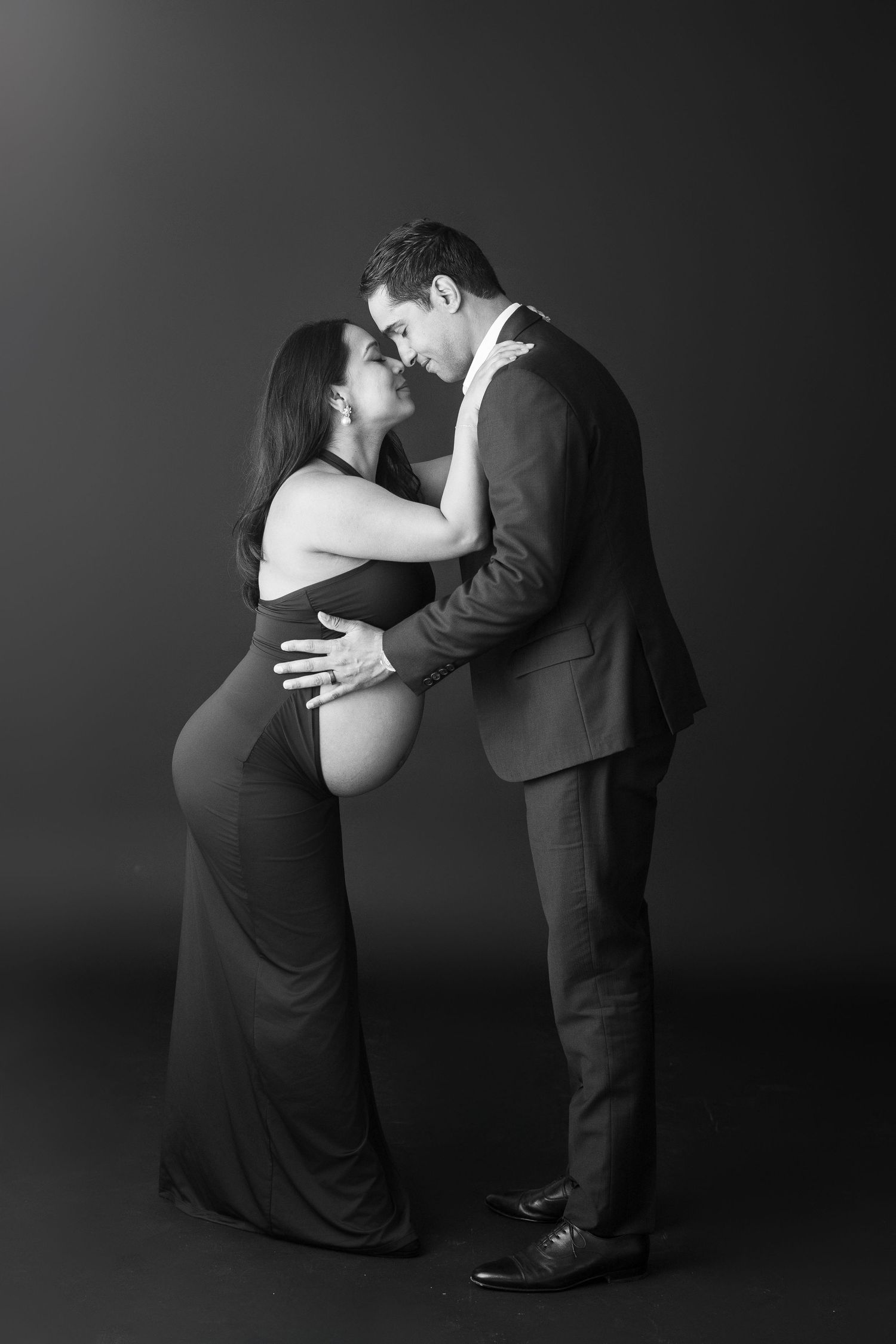 Black and white maternity photo of pregnant woman in gown embraced by suited man in professional studio.