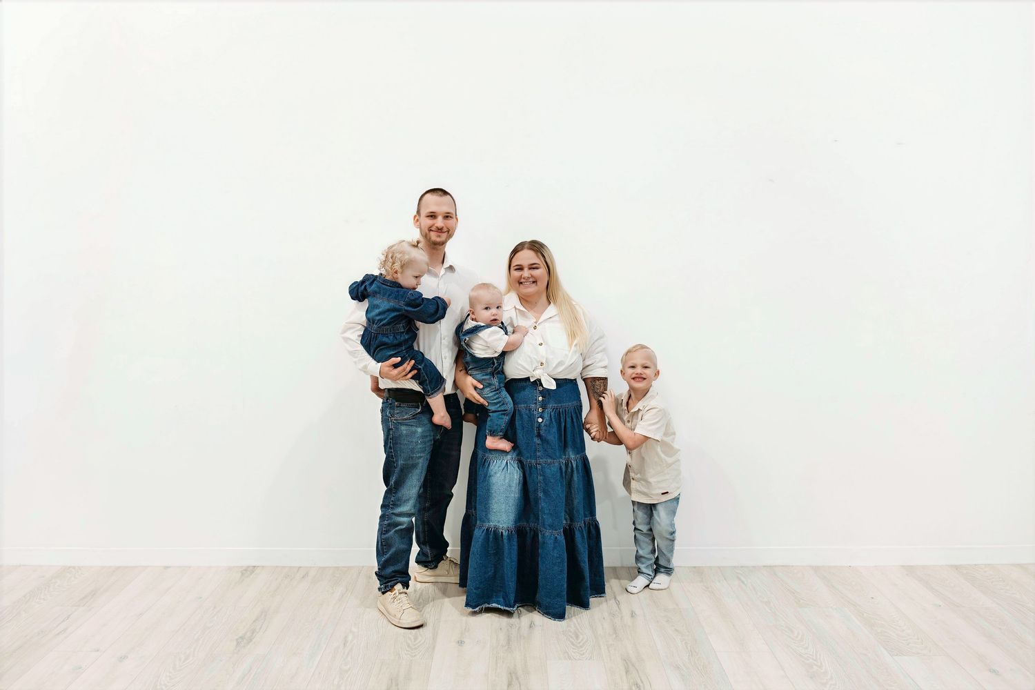 A family group in casual denim and white outfits sits together against a white background for a minimalist portrait.
