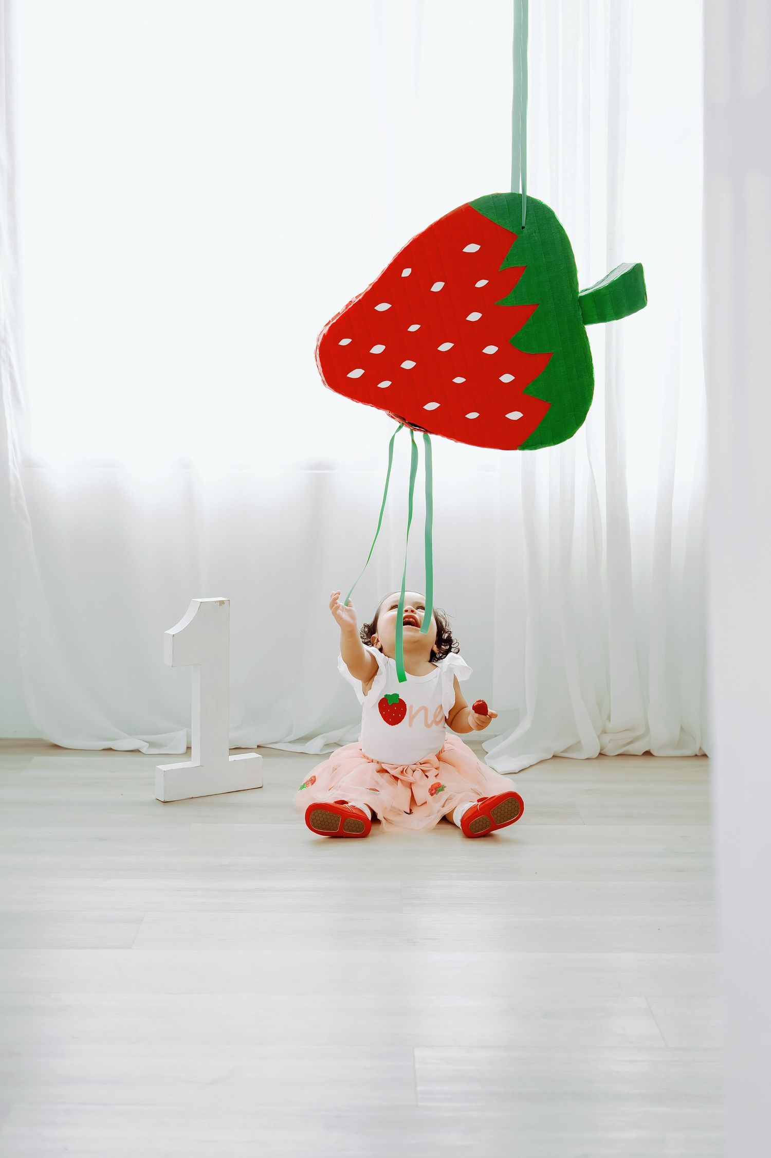 Playful studio photos with hanging strawberry decorations and white curtains in background.