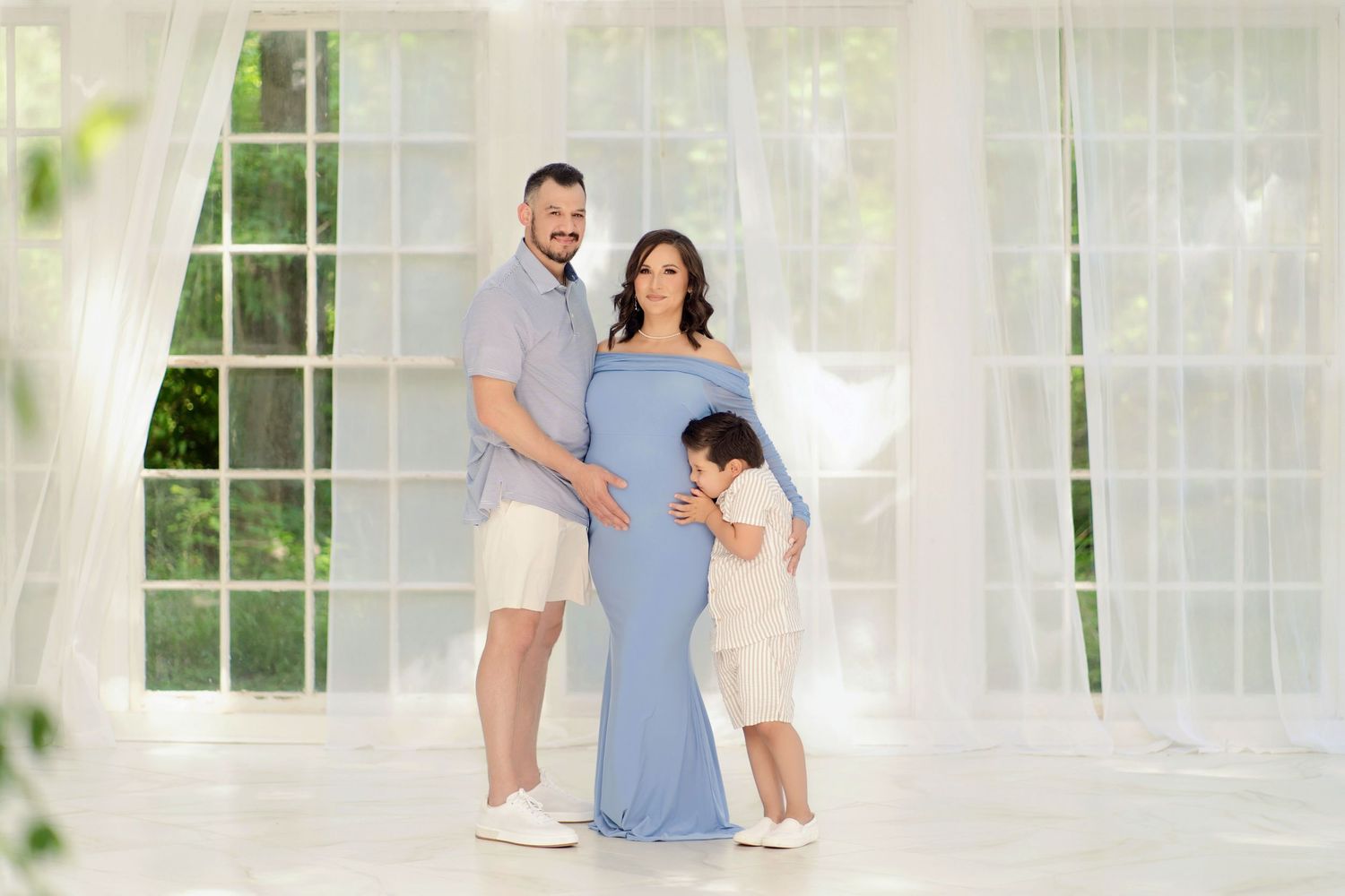 Family maternity photo with pregnant woman in blue gown, man, and child in bright white studio.