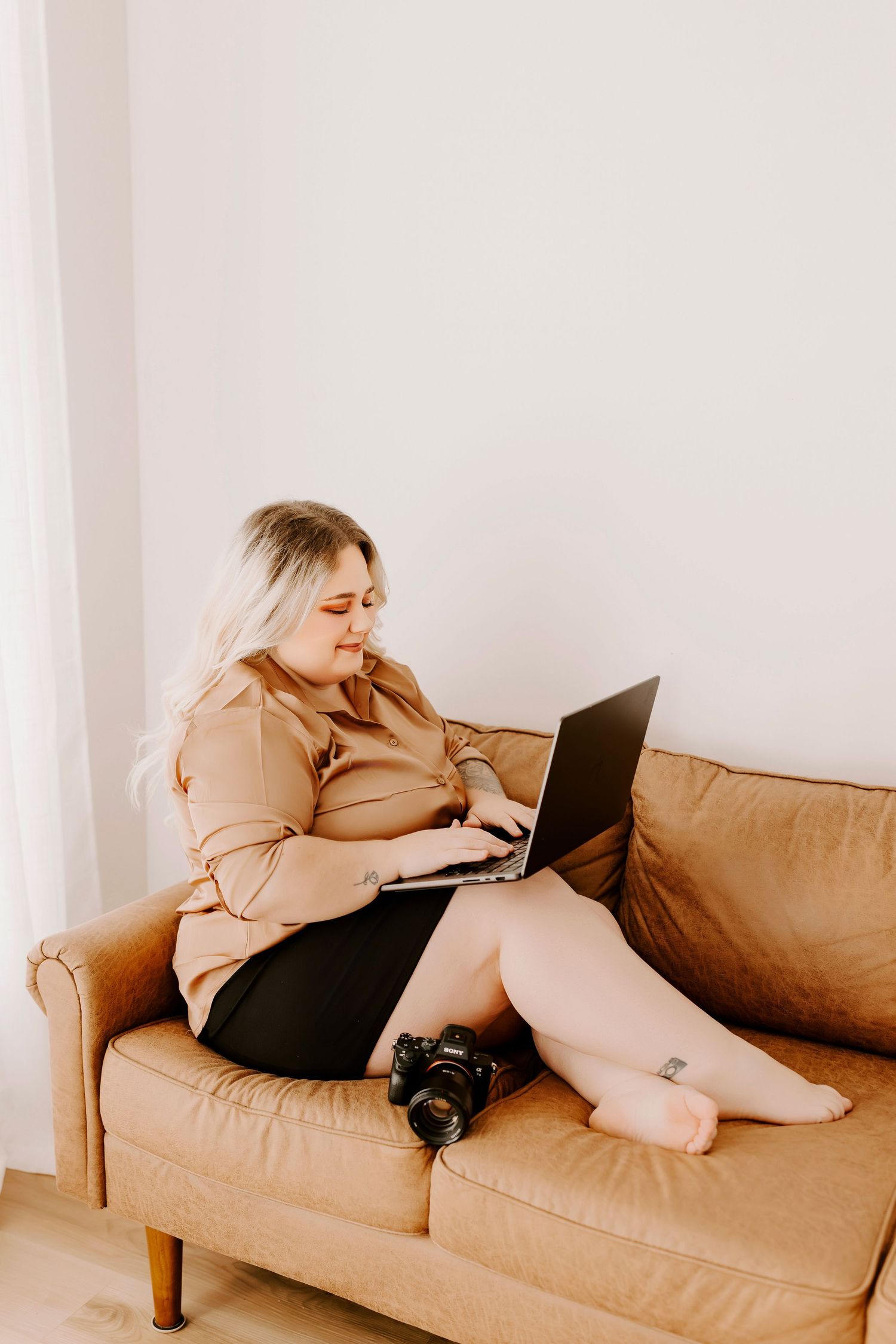 A person lounges on a tan couch while working on a laptop computer in a minimalist white room setting.