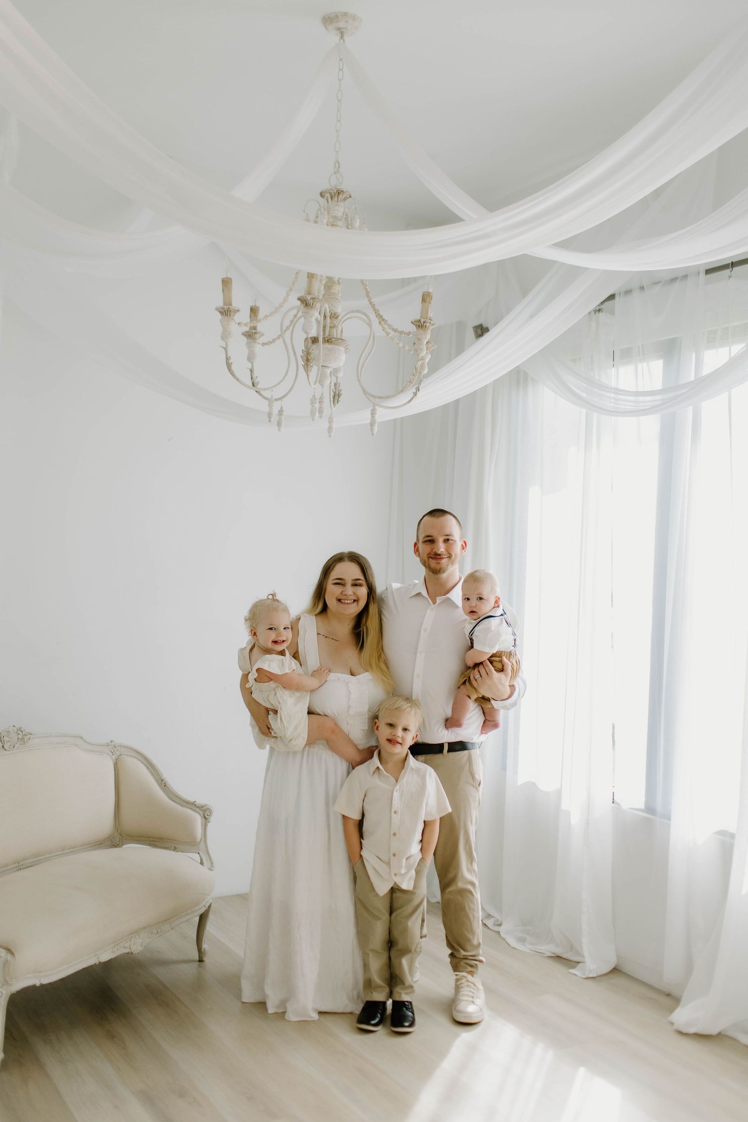 A bright white studio room with flowing sheer curtains and elegant chandelier creates a dreamy photoshoot setting.