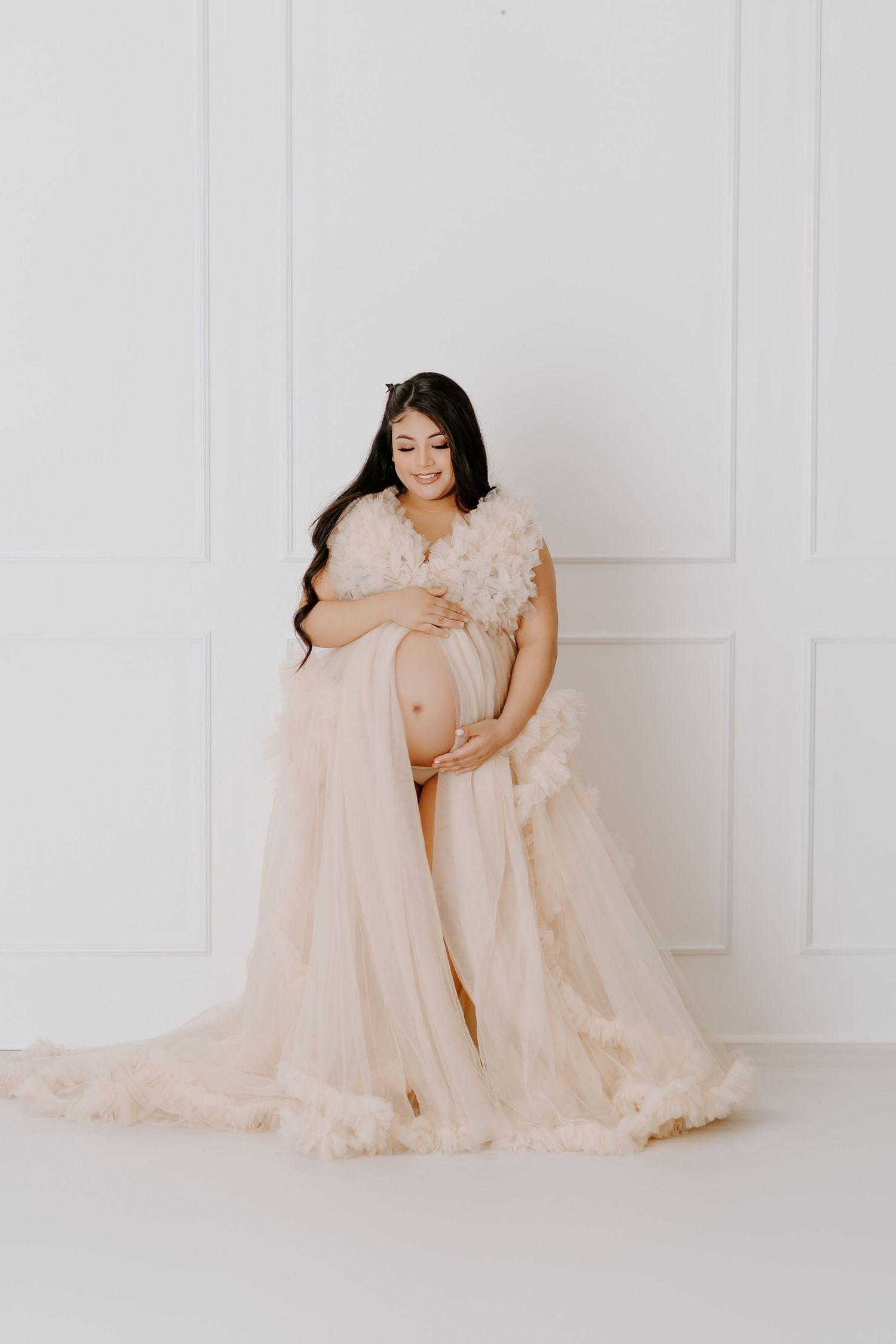 Pregnant woman in elegant blush tulle maternity gown cradling baby bump against white studio backdrop.