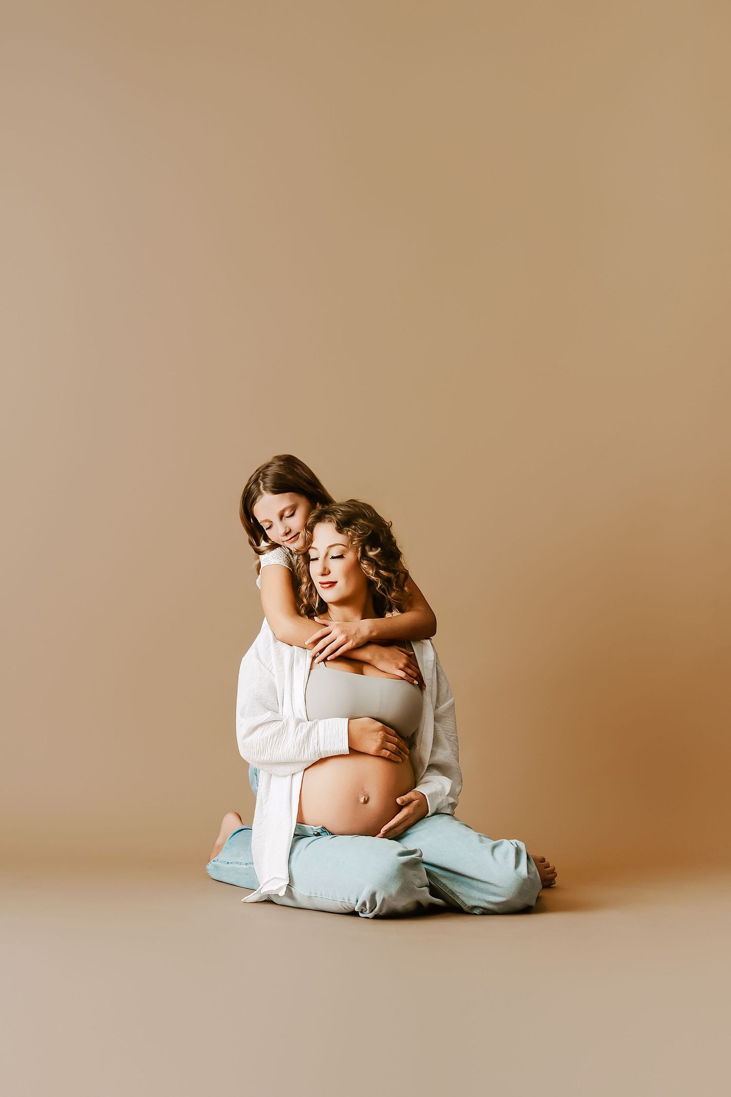 A pregnant woman sits on the floor while her young daughter hugs her from behind against a beige background.