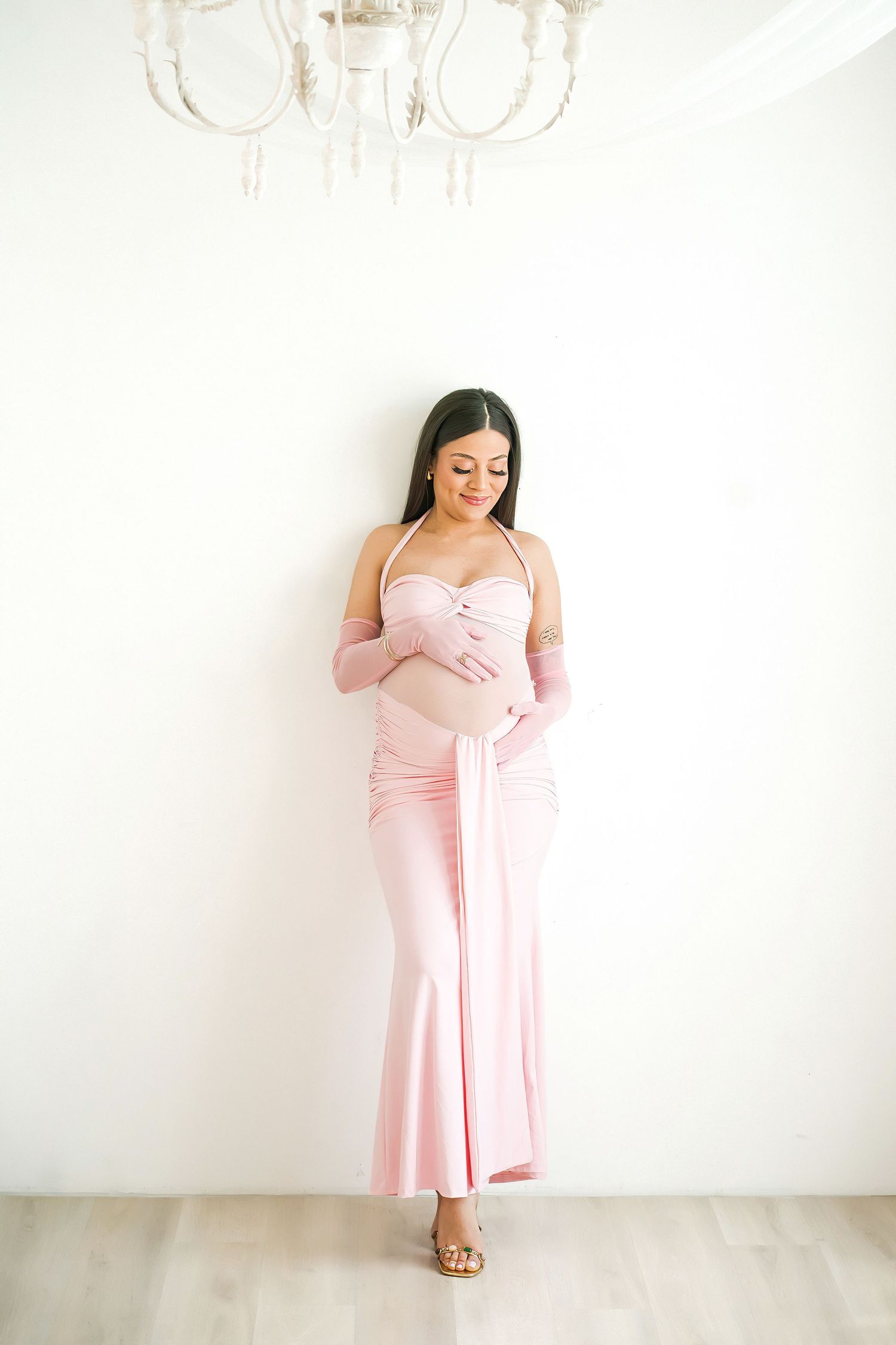 Pregnant woman in pink halter maxi dress and gloves cradling baby bump, standing against white wall.