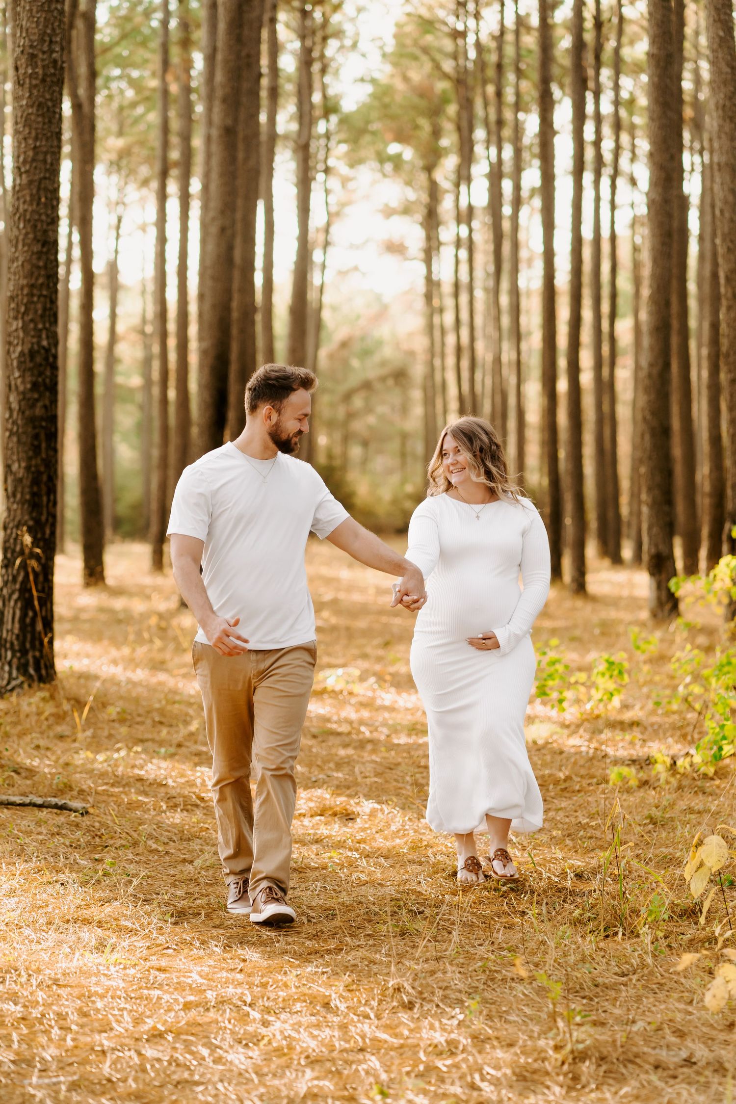 A couple in white and beige outfits walks hand in hand through a sunlit pine forest with fallen needles on the ground.