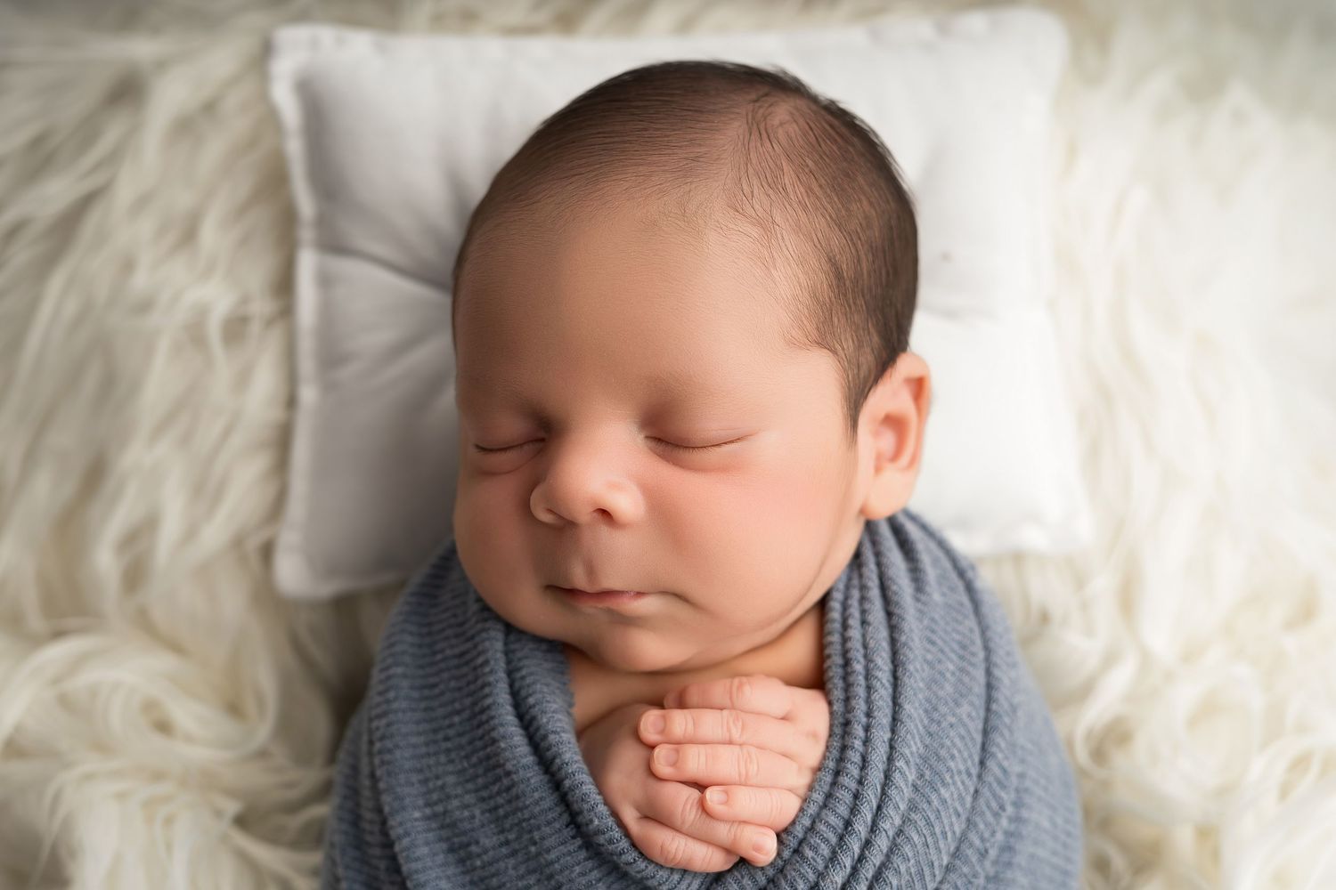 Newborn baby swaddled in blue wrap sleeping peacefully on white fluffy blanket with tiny pillow.