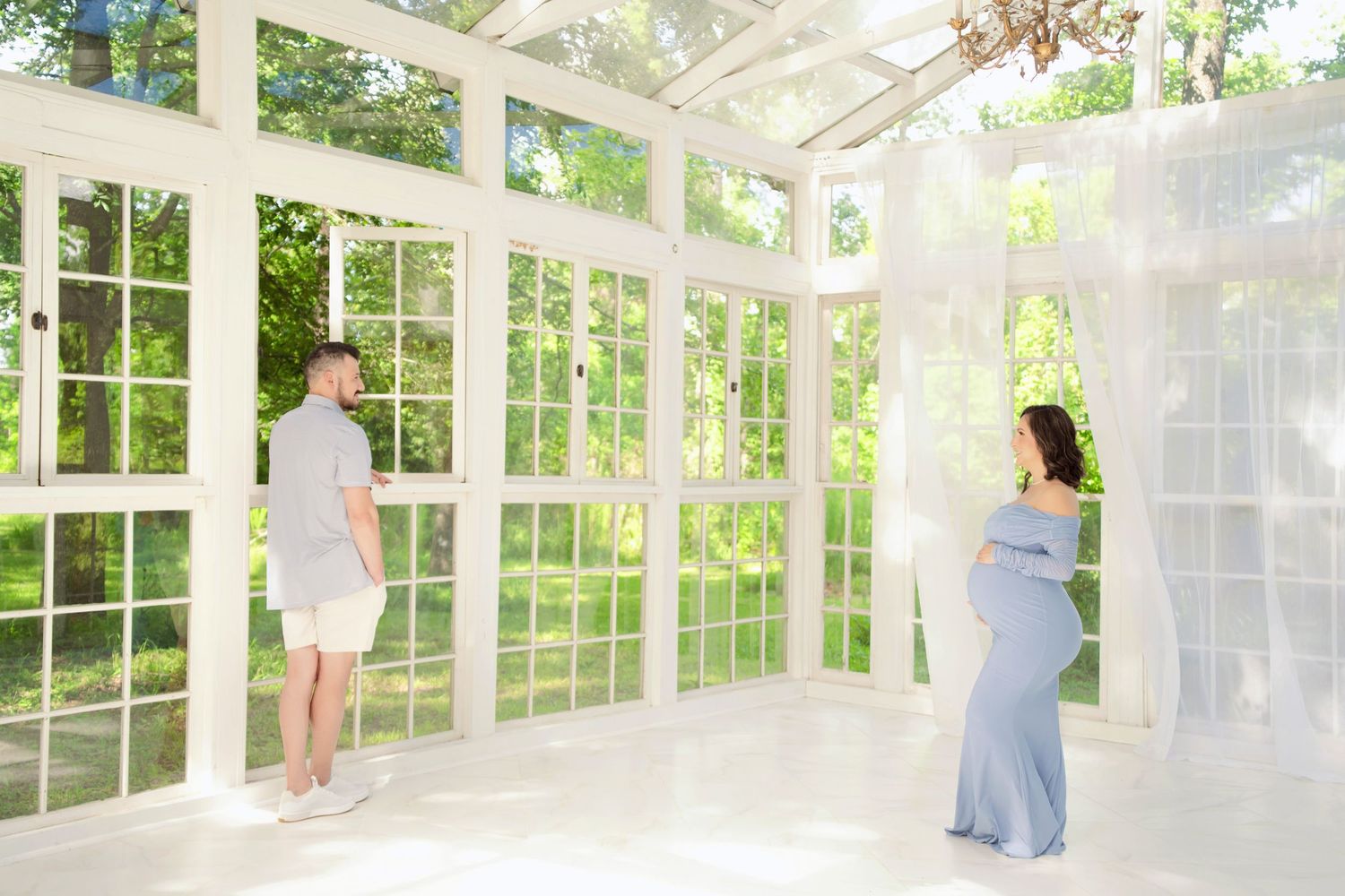 Couple in bright white sunroom studio, man in gray shirt, woman in blue maternity gown, surrounded by sheer curtains and greenery.