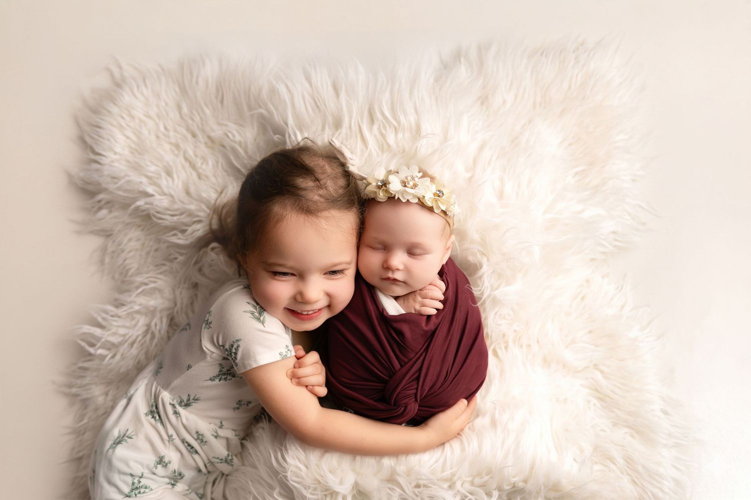 Toddler smiling and hugging swaddled newborn baby with floral headband on white fluffy blanket.