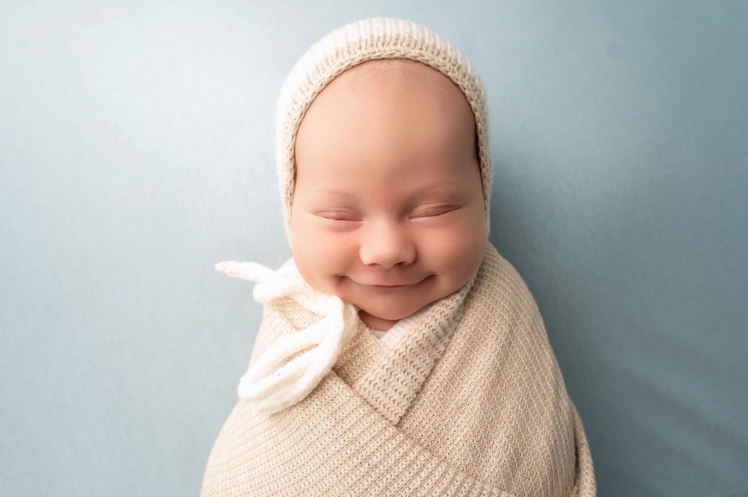 Smiling newborn baby wrapped in cream knit swaddle blanket wearing matching bonnet on soft blue background.