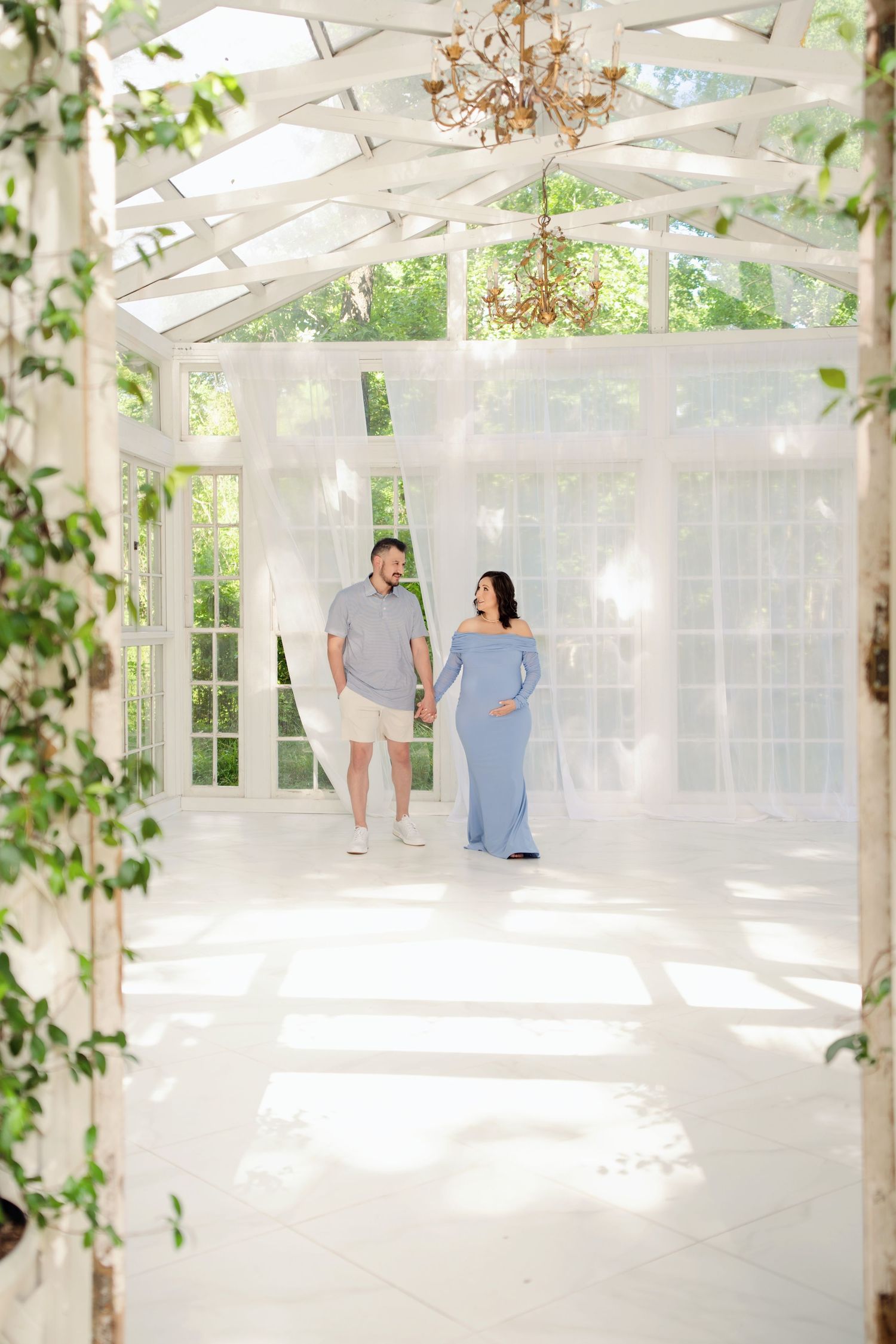 Couple holding hands in a bright white greenhouse conservatory, woman in blue maternity gown, man in gray polo and white shorts.