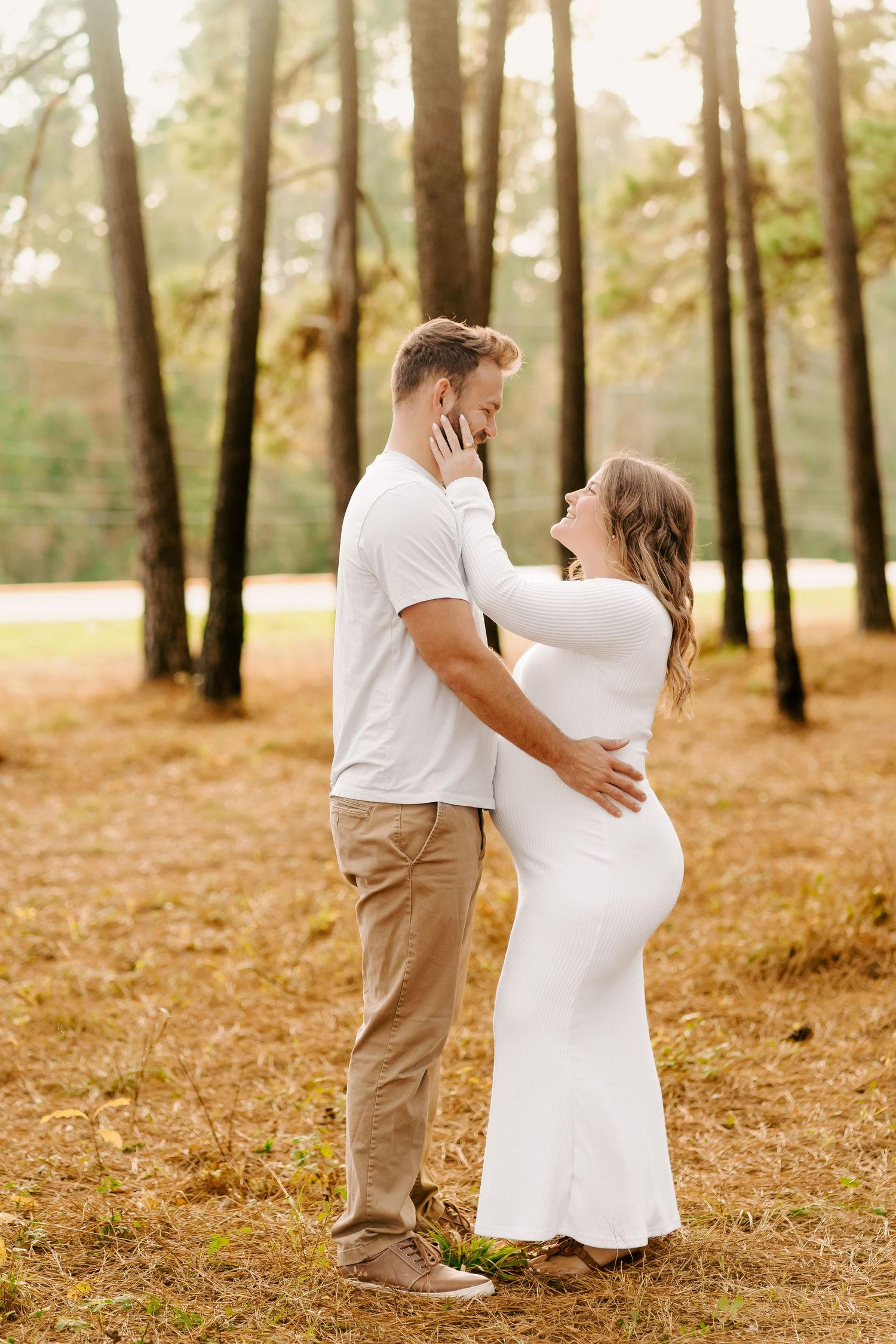 Couple in white and khaki embraces during maternity photoshoot in a sunlit pine forest.