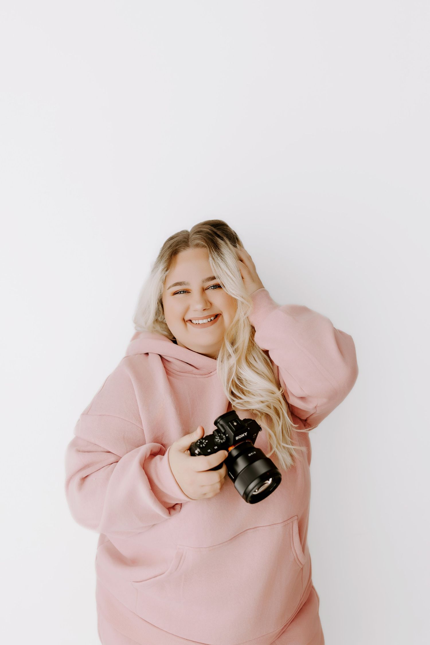 Person wearing a cozy pink hoodie holds a camera while posing against a white background.