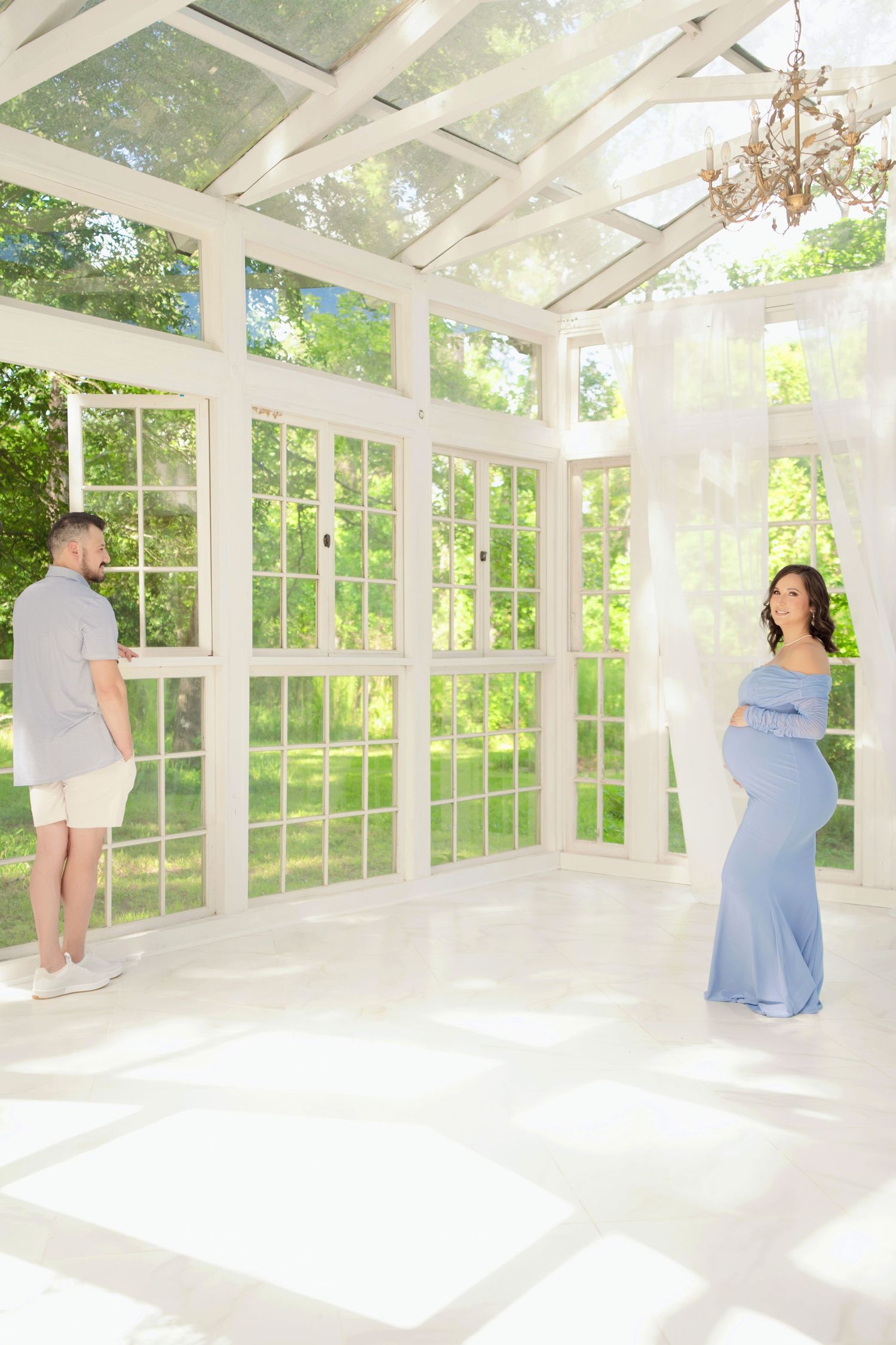 Pregnant woman in blue gown with partner in bright white sunroom greenhouse studio for maternity photo session.