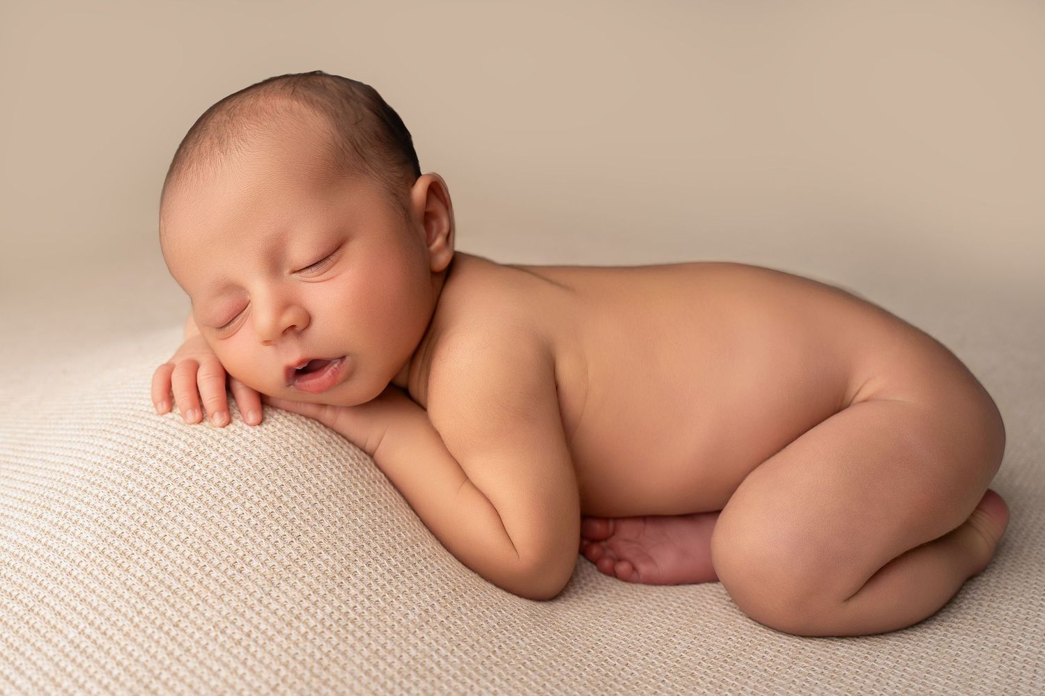 A peaceful newborn baby sleeping curled up on a soft cream blanket in a classic posed newborn photography style.