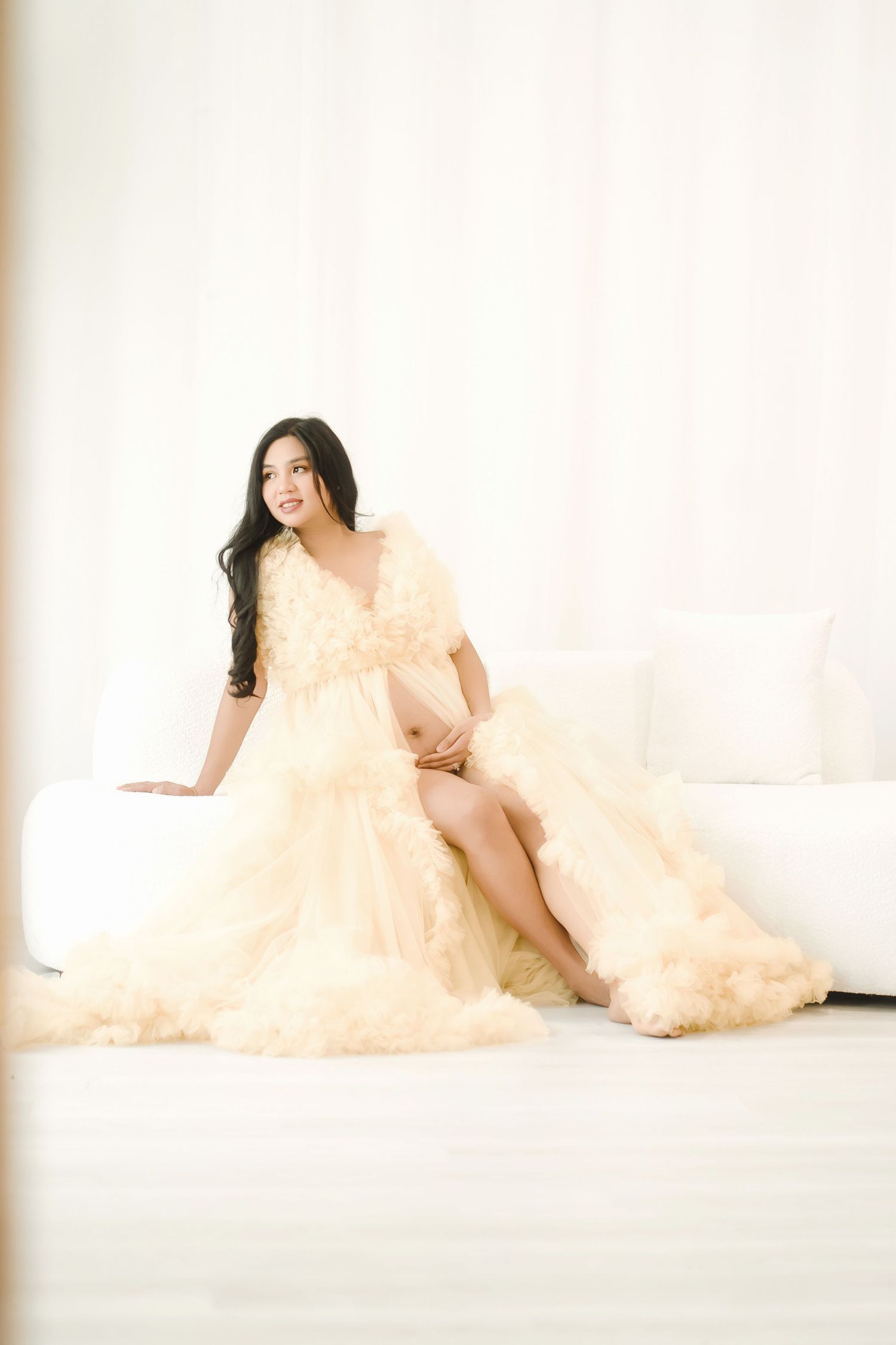 Pregnant woman in elegant cream tulle maternity gown sitting gracefully on white sofa in bright airy studio.