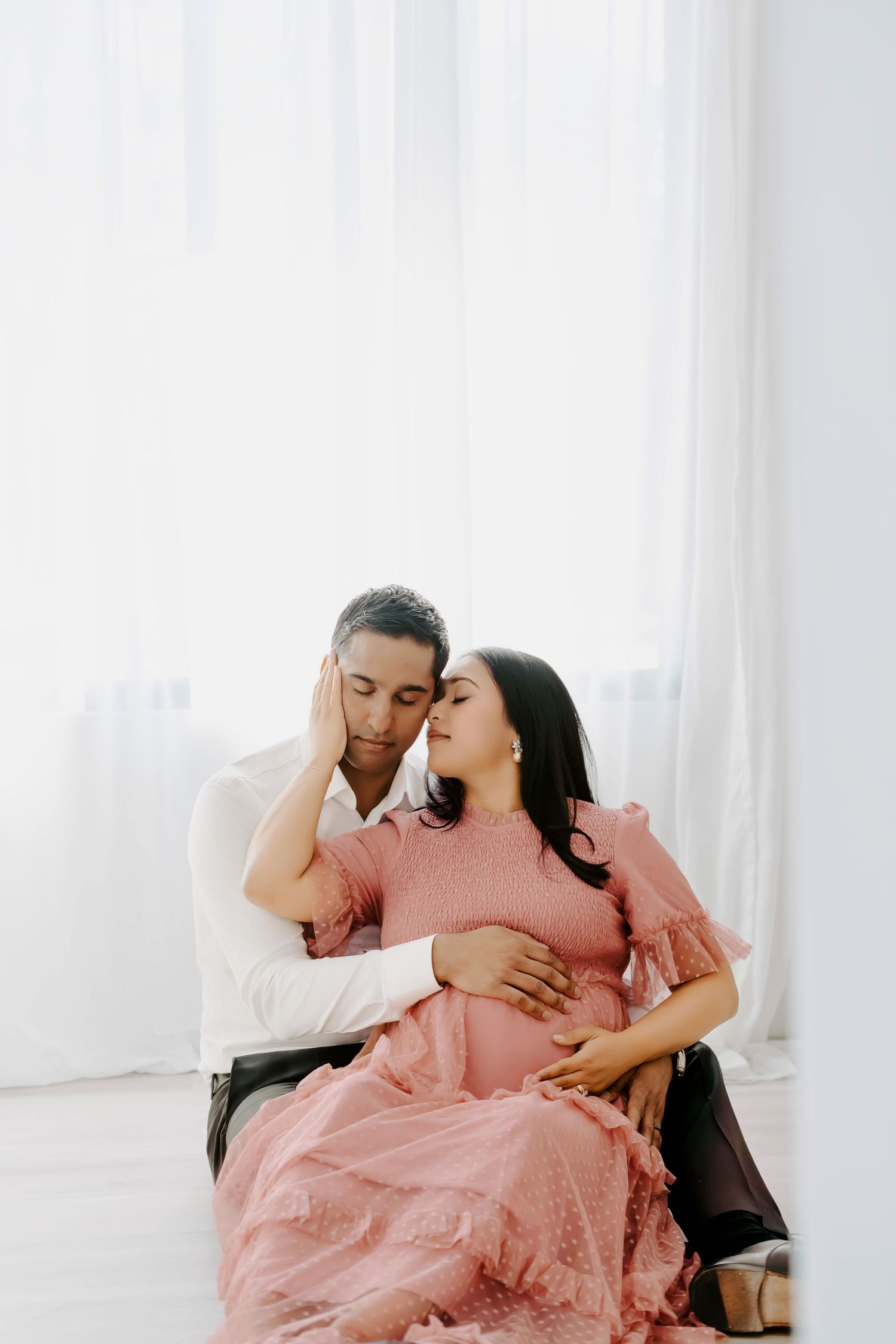 A couple shares a tender moment, the pregnant woman in a pink dress cradled by her partner in a bright white room.