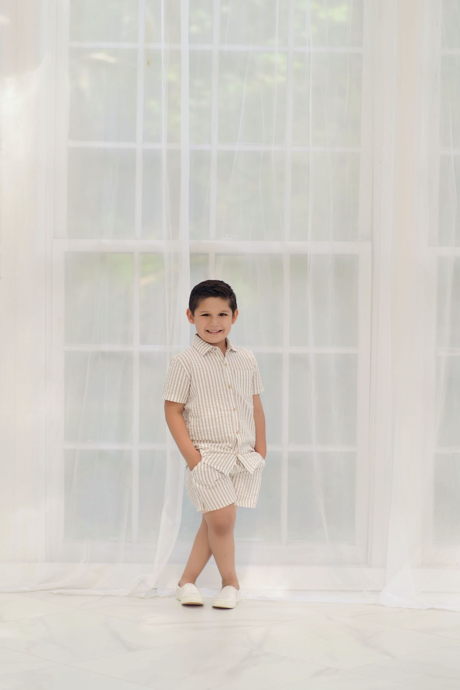 Young boy in striped linen shirt and shorts set, smiling confidently in a bright white studio setting.