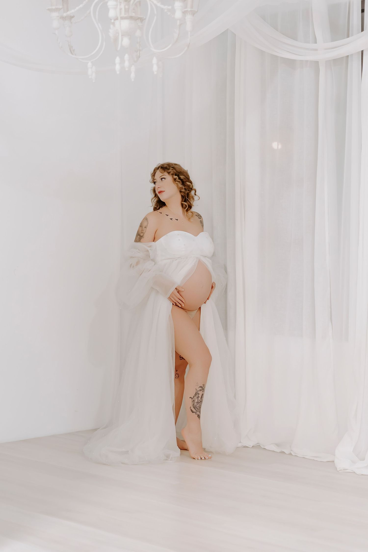 Pregnant woman in white tulle gown holding baby bump in bright white studio with chandelier and sheer curtains.
