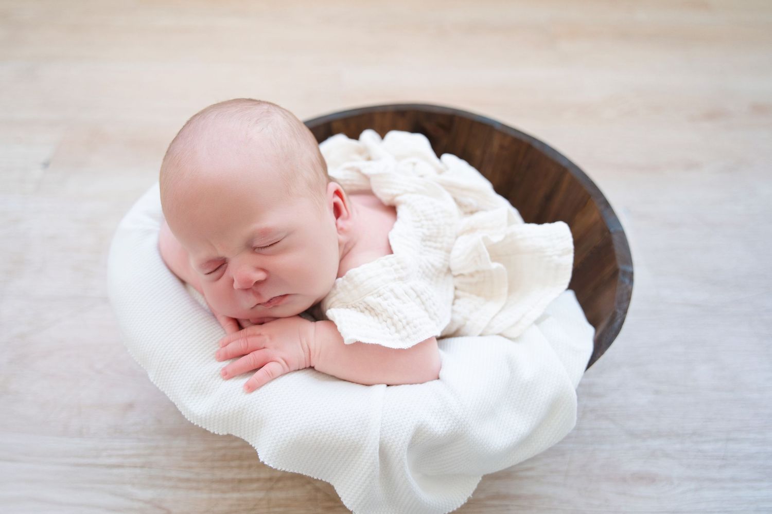 A sleeping newborn baby wrapped in white fabric rests peacefully in a wooden bowl.