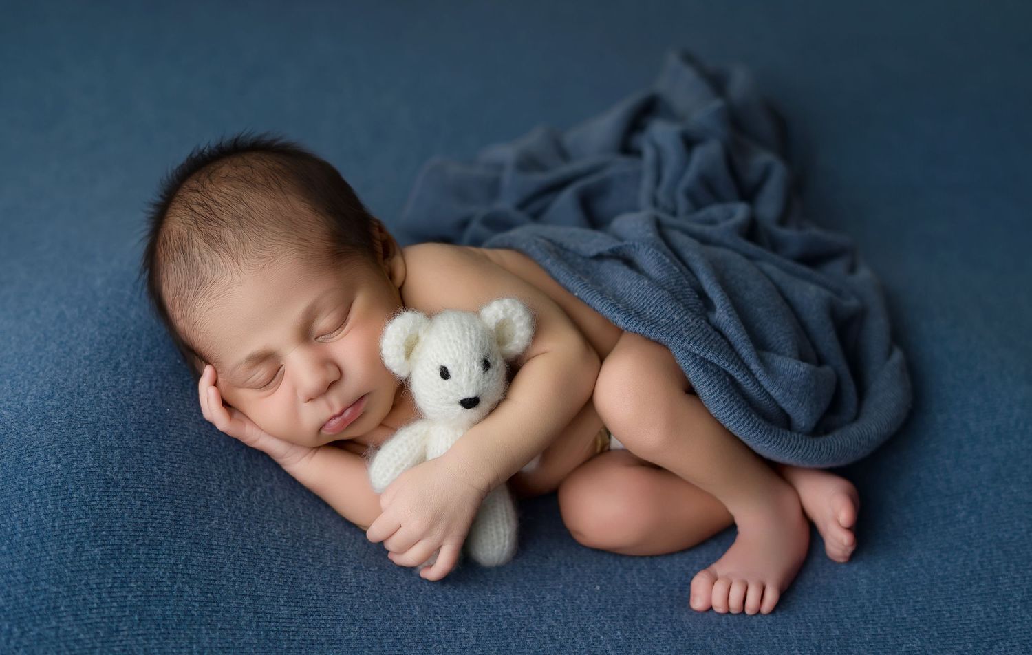 Peaceful newborn baby sleeping curled up with a white teddy bear, wrapped in a soft blue blanket.