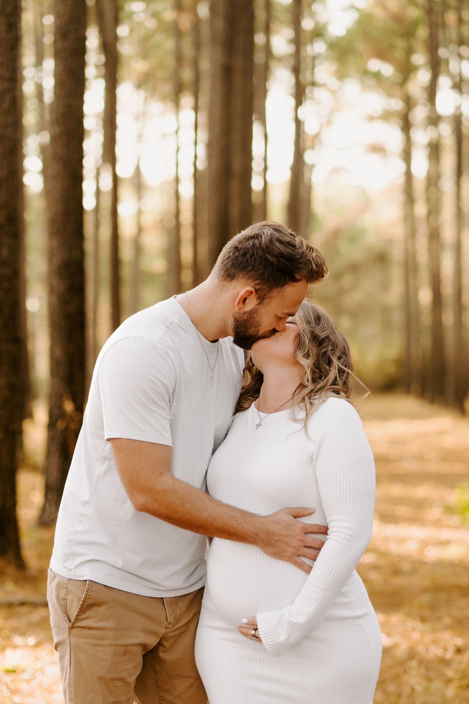 Romantic couple in white outfits embrace tenderly in a sunlit forest with autumn leaves on the ground.