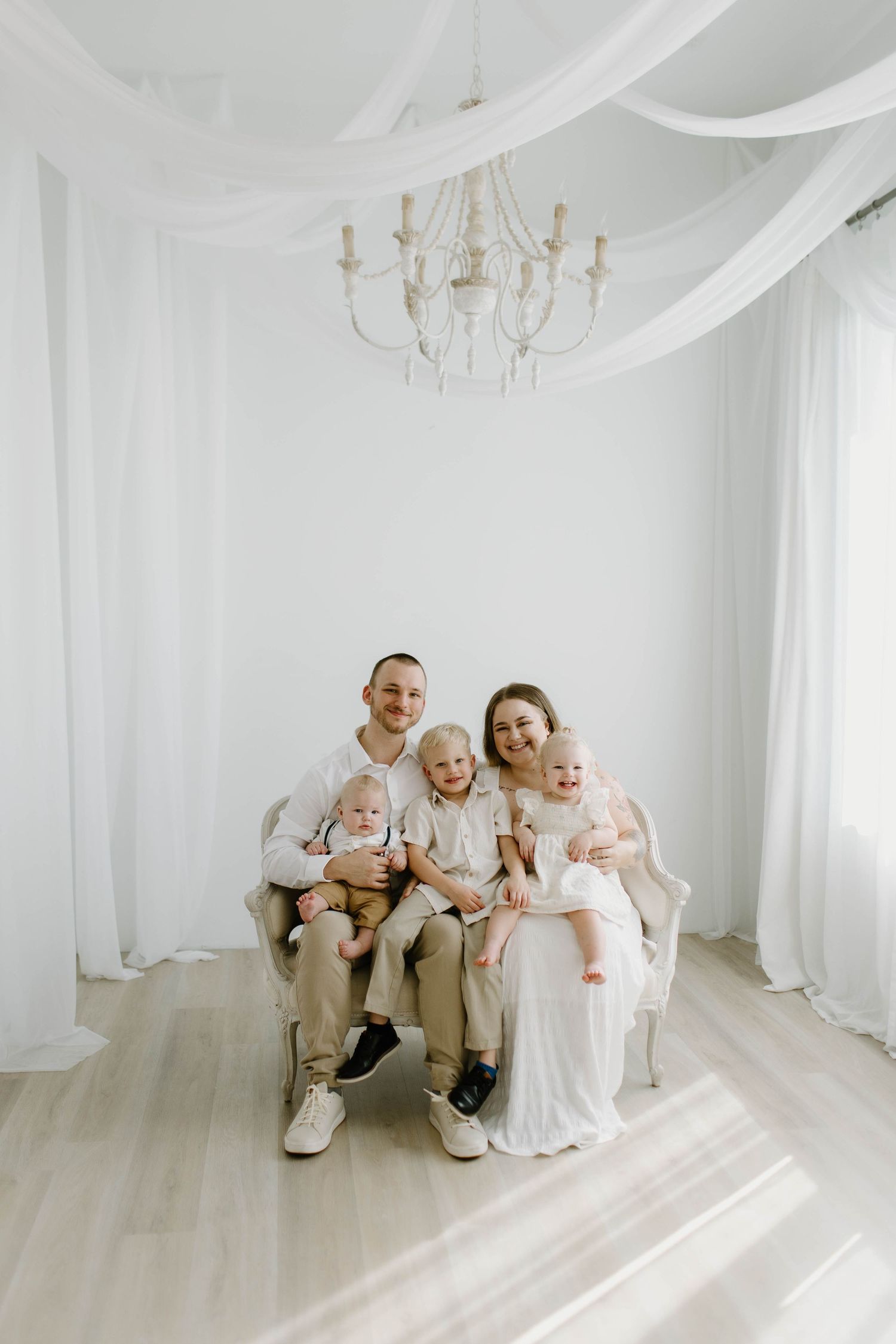 A family group sits together in an elegant all-white studio room with flowing fabric drapes and a chandelier.
