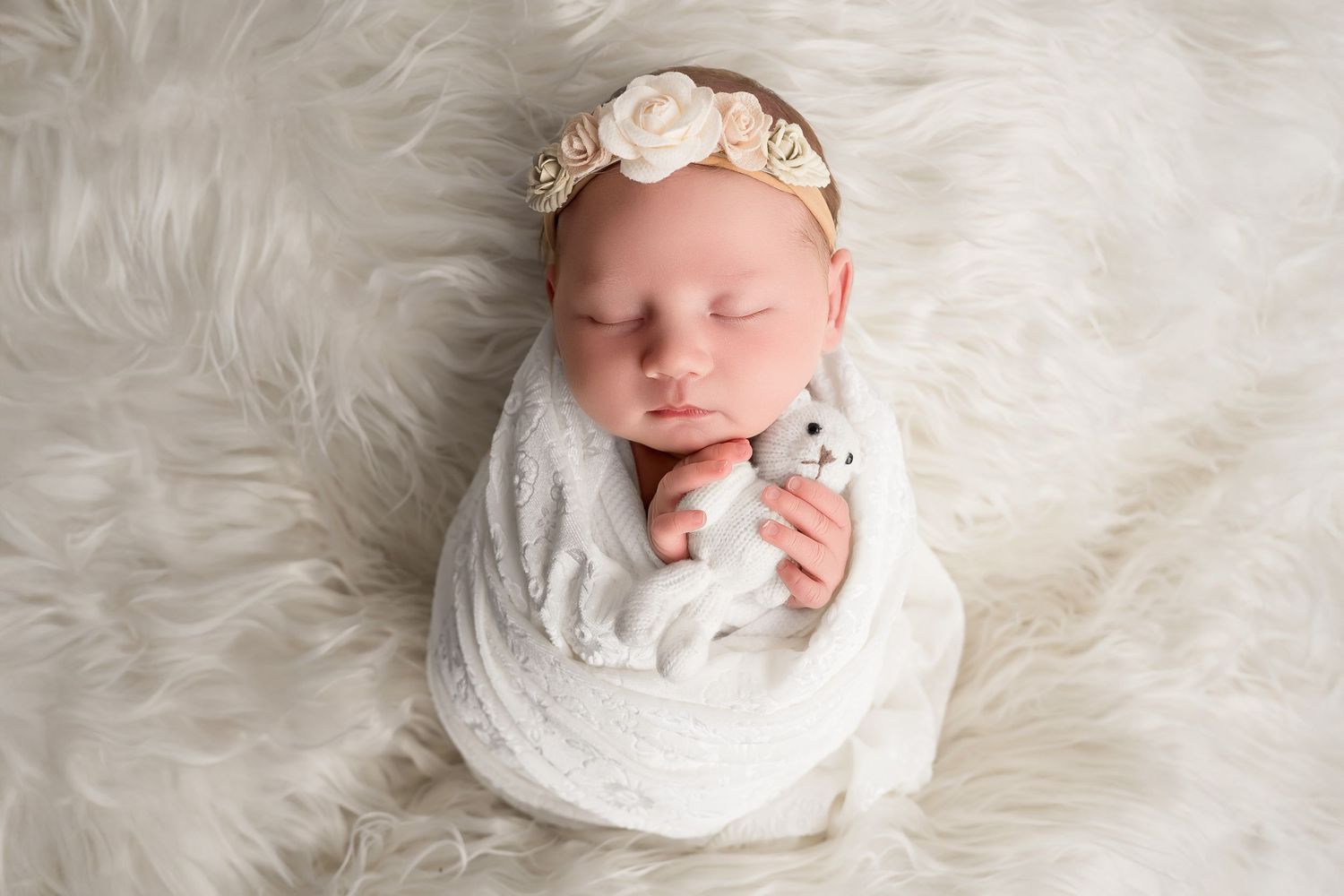 Sleeping newborn baby swaddled in white lace wrap with floral headband holding tiny teddy bear on fluffy white fur backdrop.