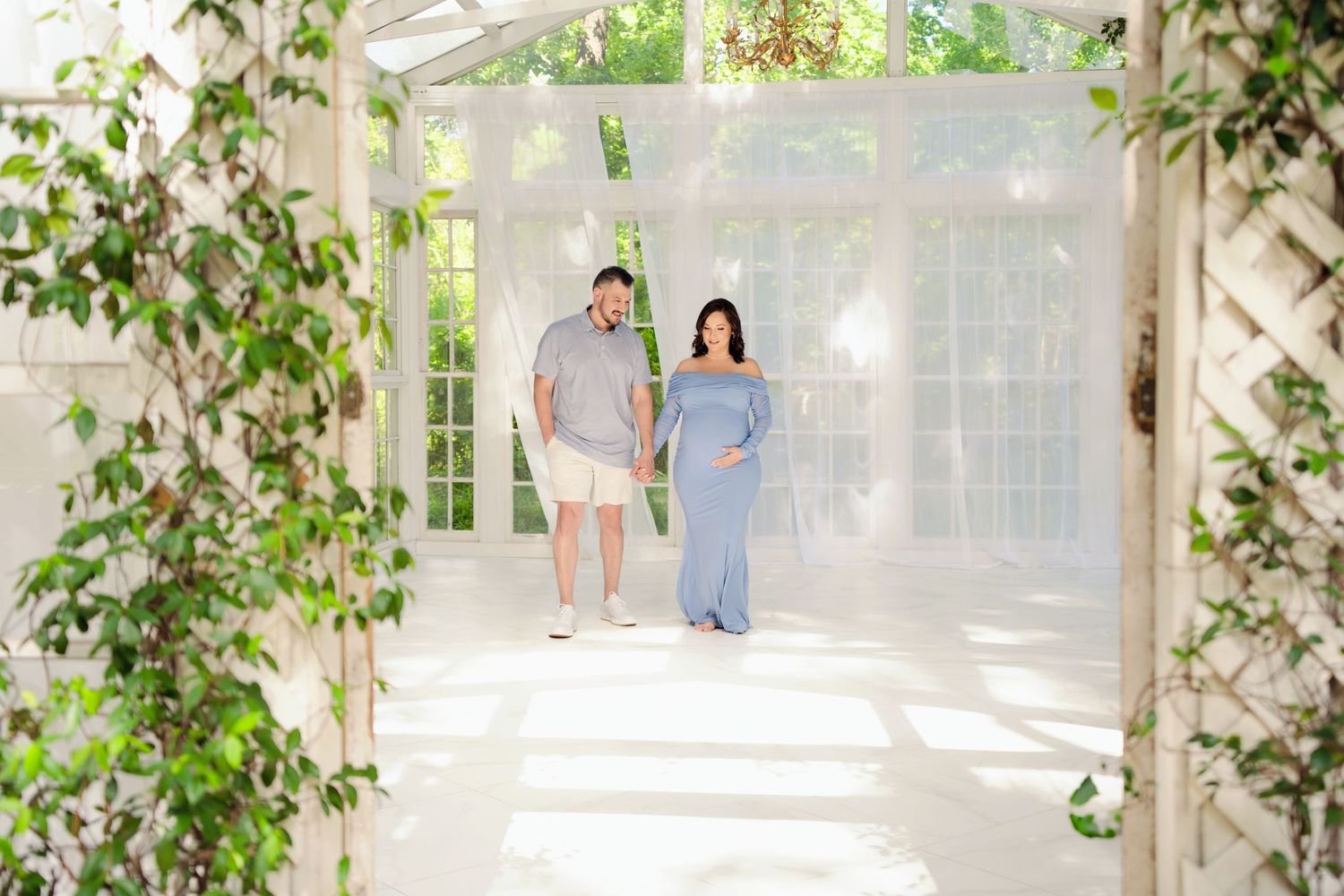 Couple maternity shoot, pregnant woman in blue gown holding hands with man in white garden studio.
