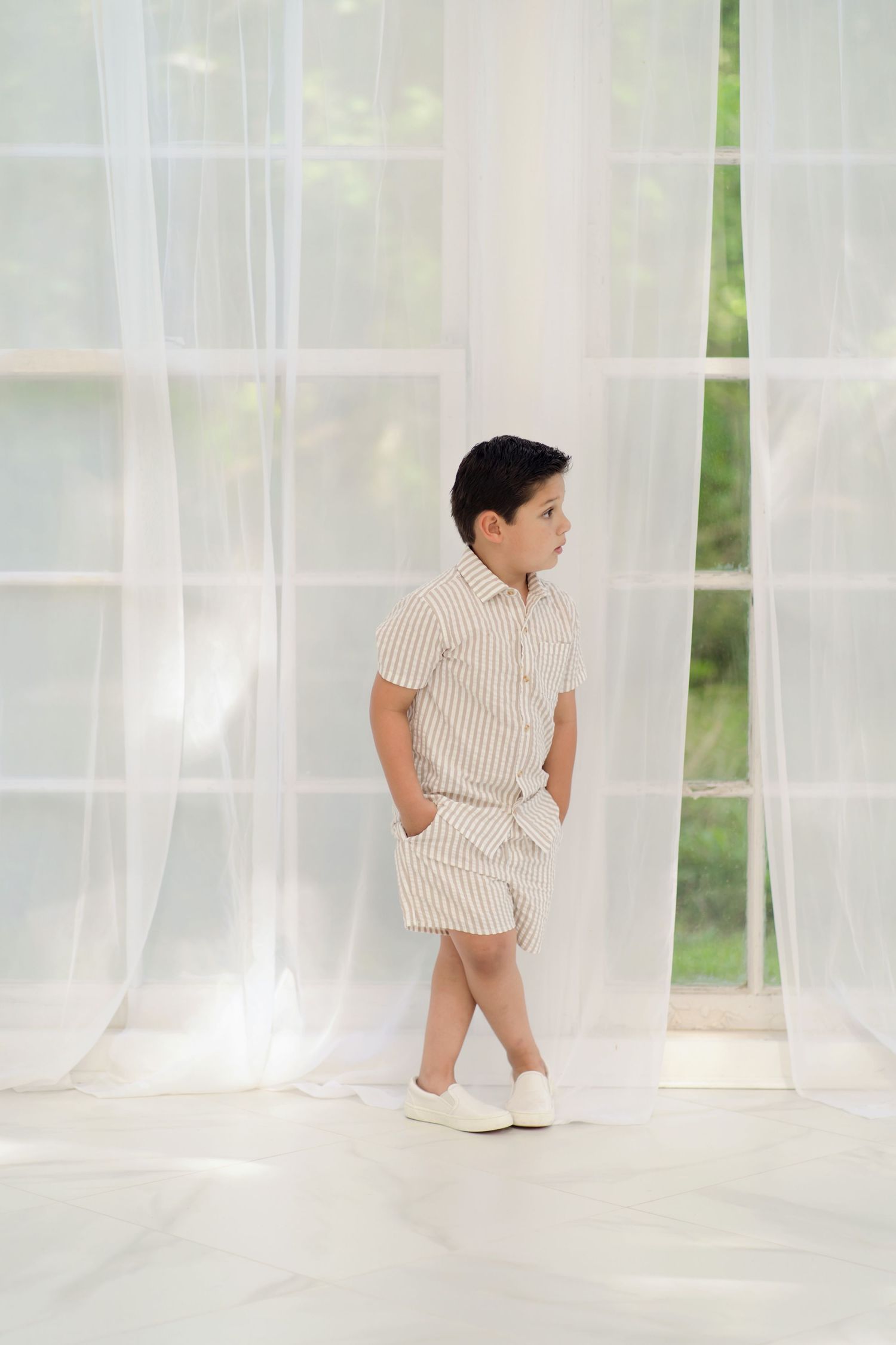 Young boy in striped shirt and shorts stands by sheer white curtains, looking sideways in a bright room.