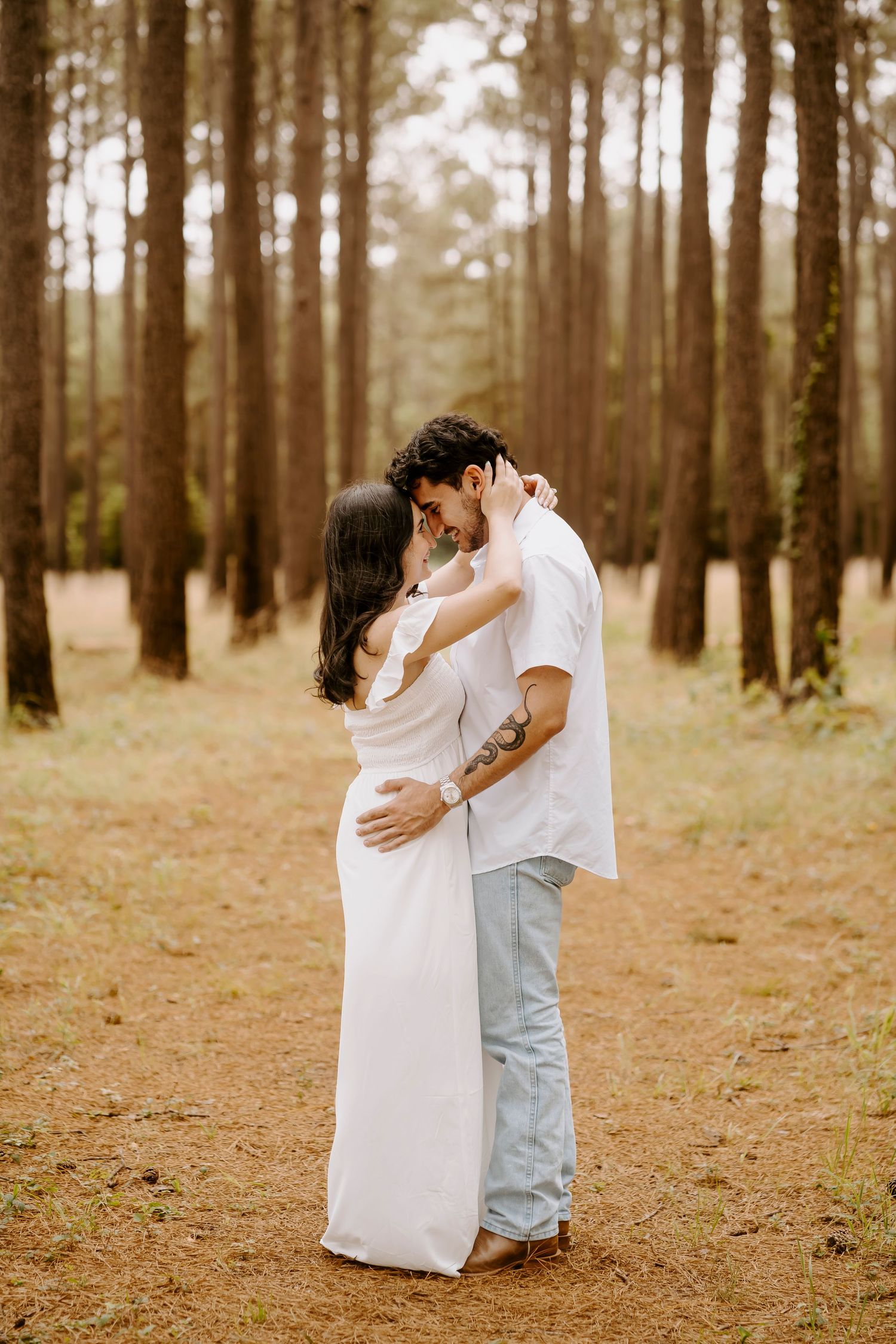 Couple embracing and almost kissing in a pine forest, woman in white dress, man in white shirt and jeans.