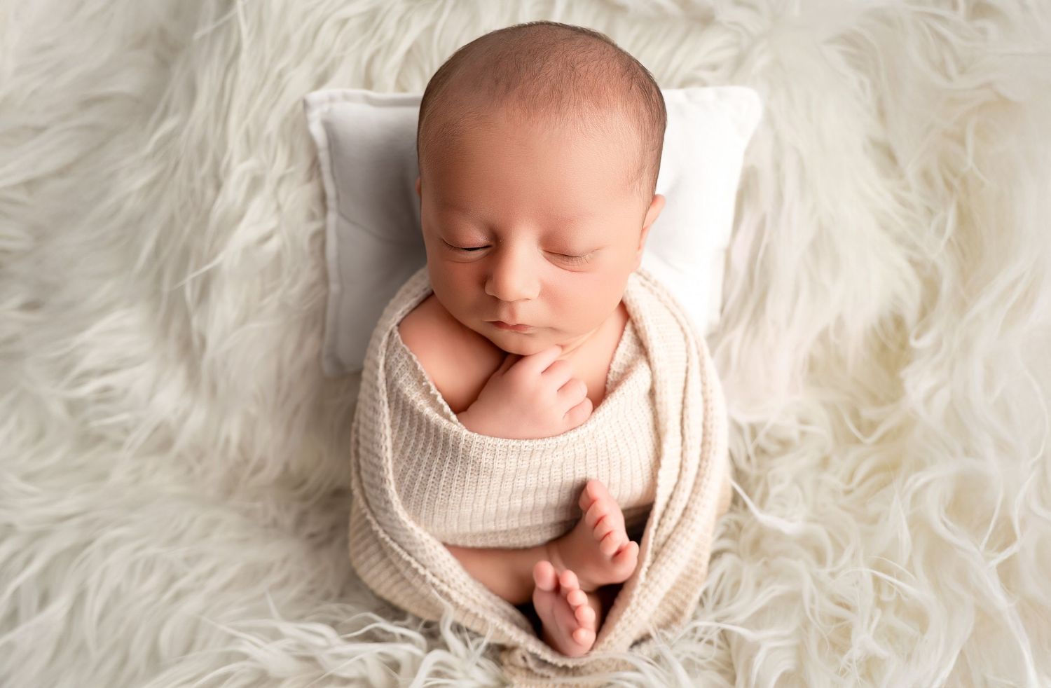 Peaceful newborn baby swaddled in cream knit wrap sleeping on fluffy white fur blanket with tiny pillow.