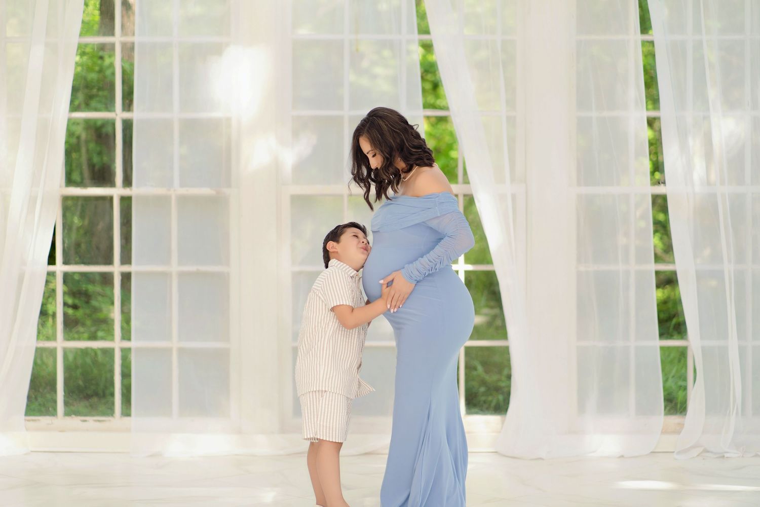 A young boy tenderly touches a pregnant woman's baby bump in a bright, airy studio during a maternity photo session.