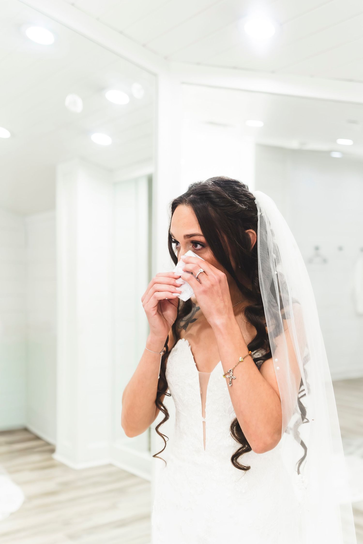 Emotional bride in white wedding dress and veil dabbing tears with tissue in bright bridal suite.