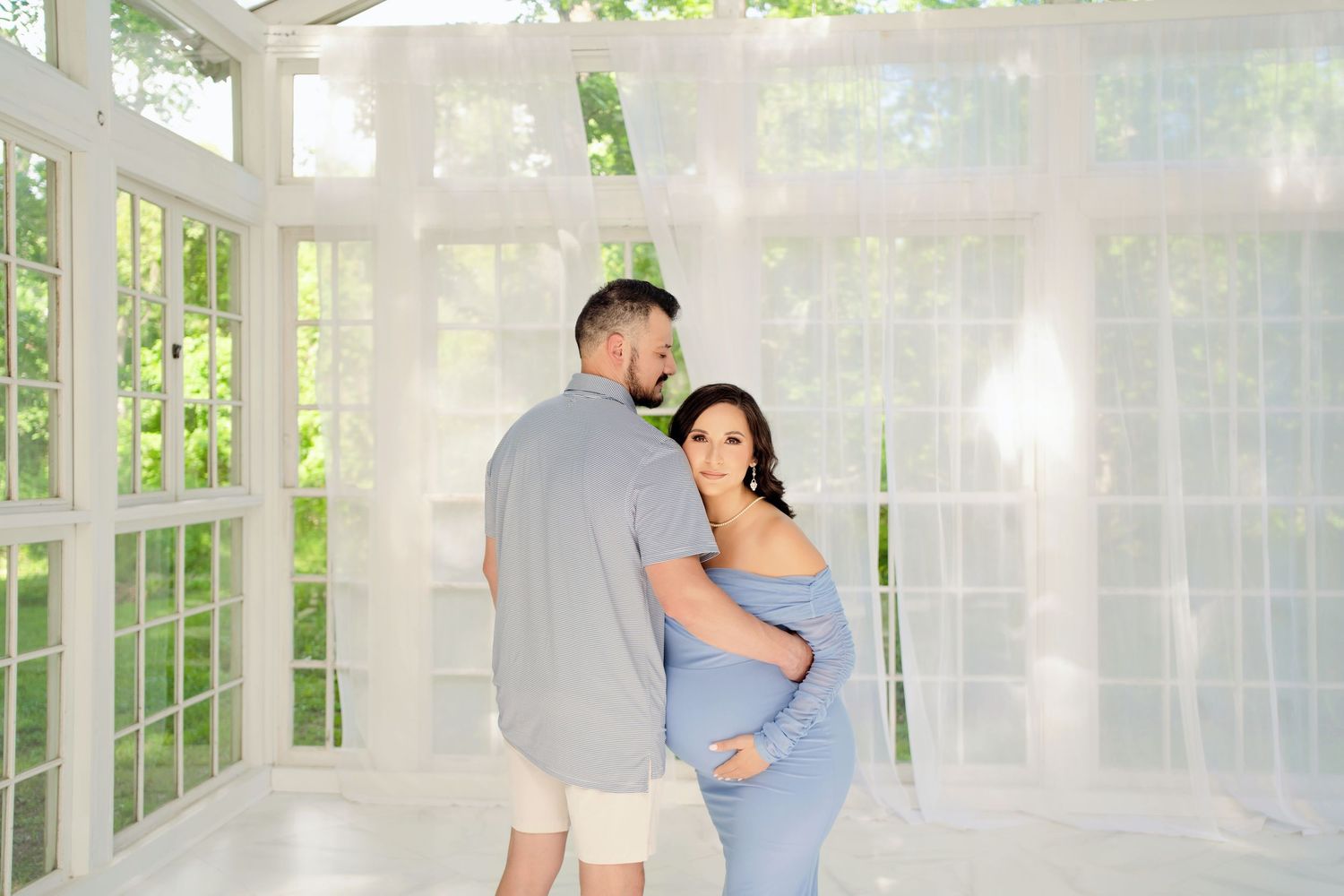 Couple in matching blue outfits posing in a bright white studio with sheer curtains, woman in maternity dress.