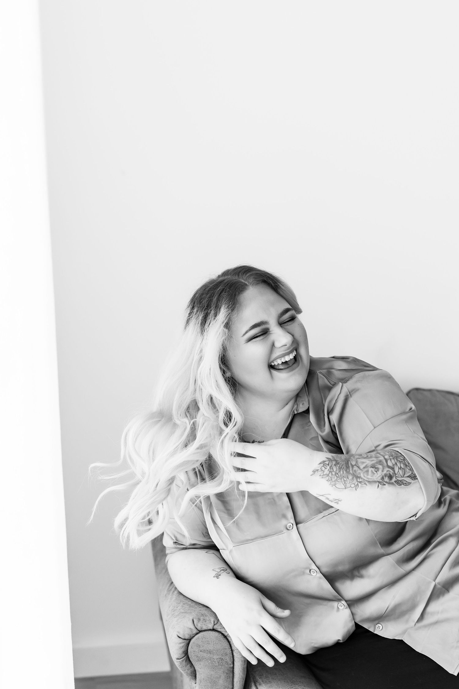Black and white photo of a laughing woman with long blonde hair and tattoos sitting in a chair.