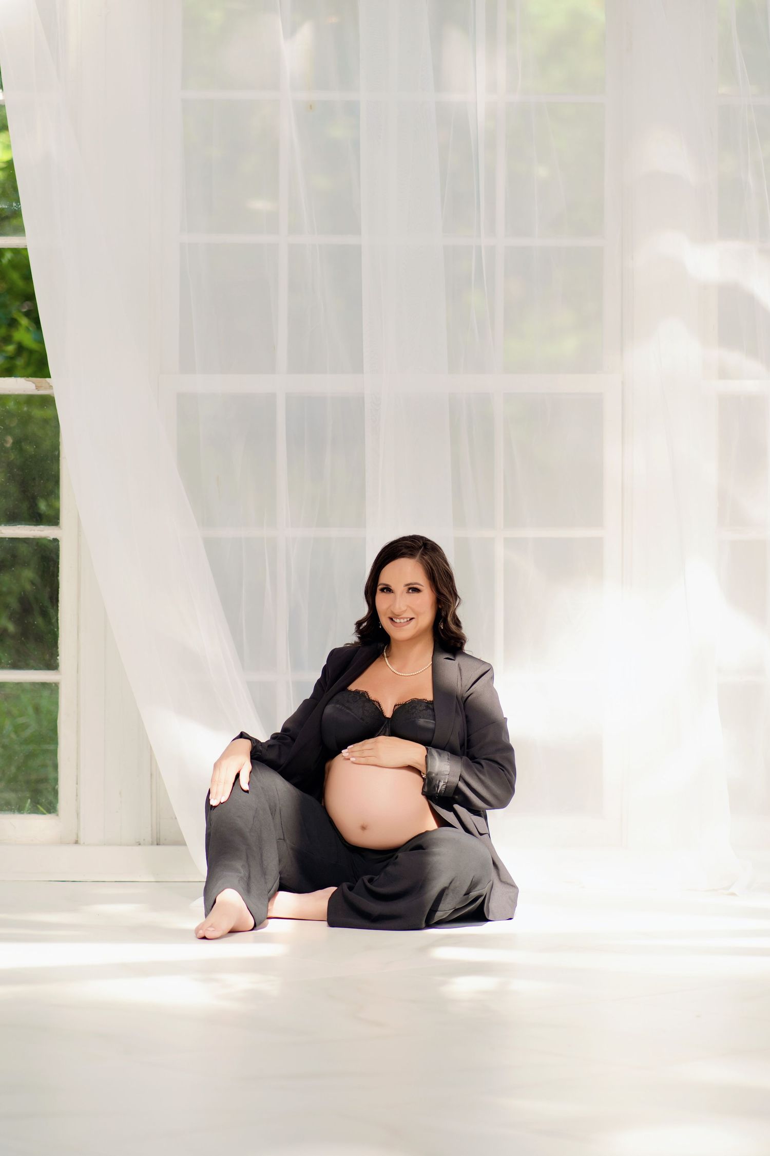 Pregnant woman in gray blazer sitting on white floor, cradling baby bump, smiling near sheer curtains.