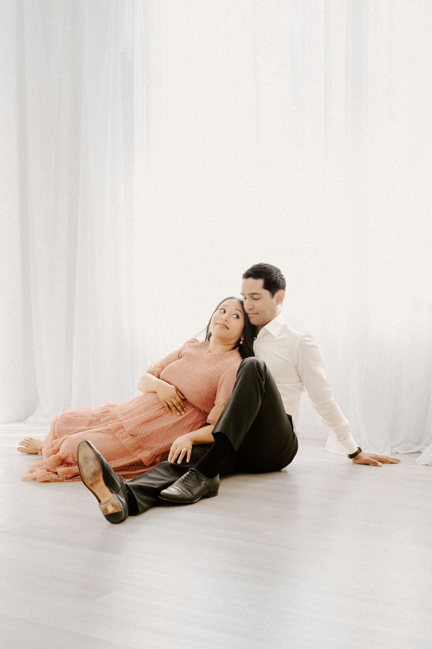 Pregnant couple sitting on white studio floor, woman in pink dress leaning against man in white shirt, maternity photo.