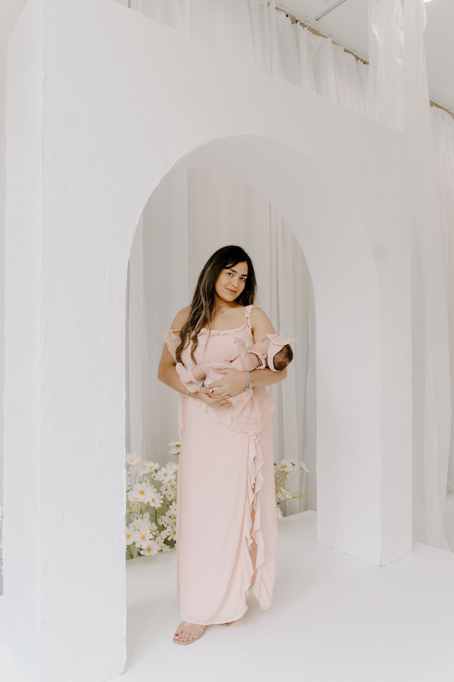 Woman in blush pink ruffle maxi dress holding newborn baby in white arch studio setting.