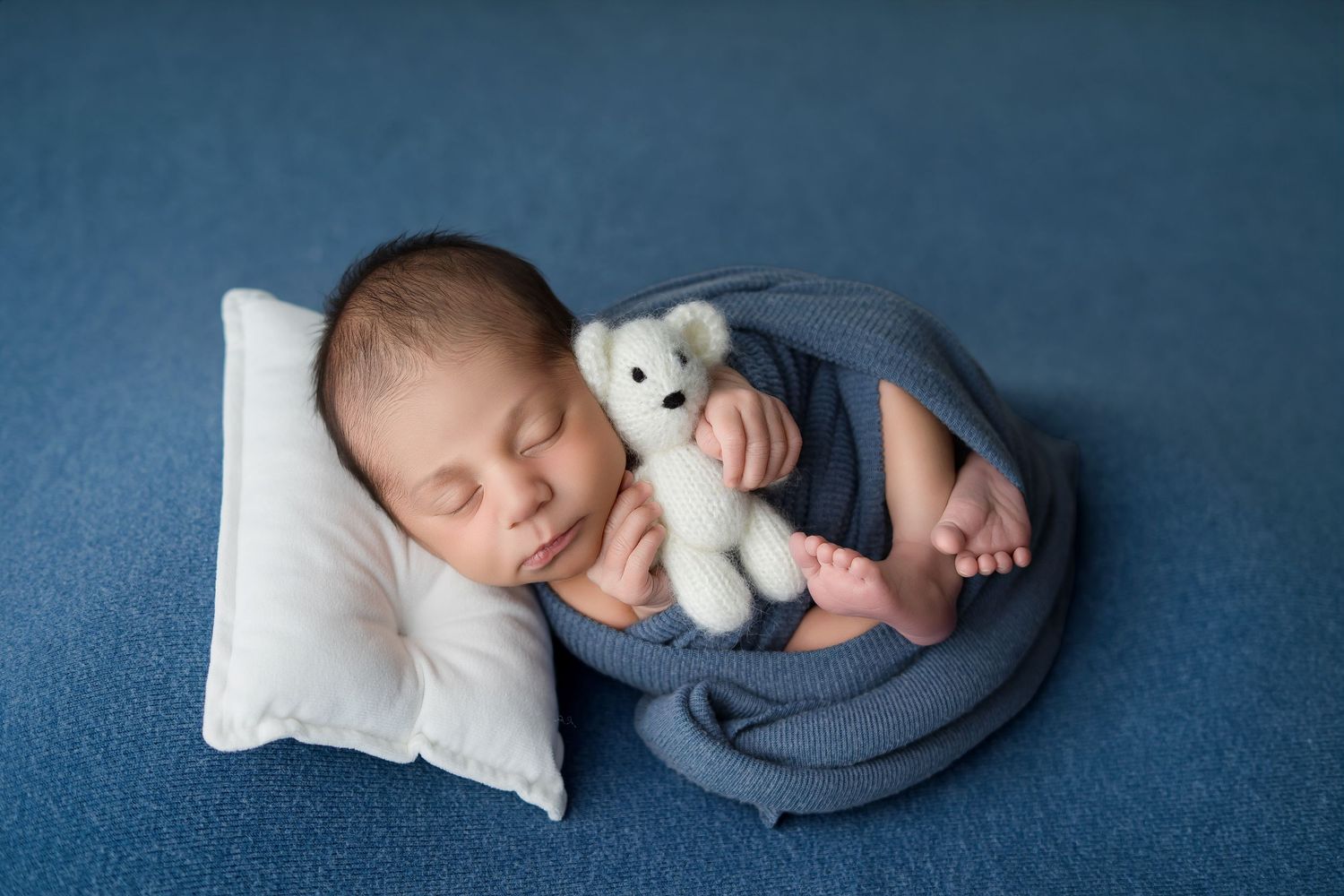 Newborn baby sleeping wrapped in blue fabric holding a white teddy bear on a blue background.