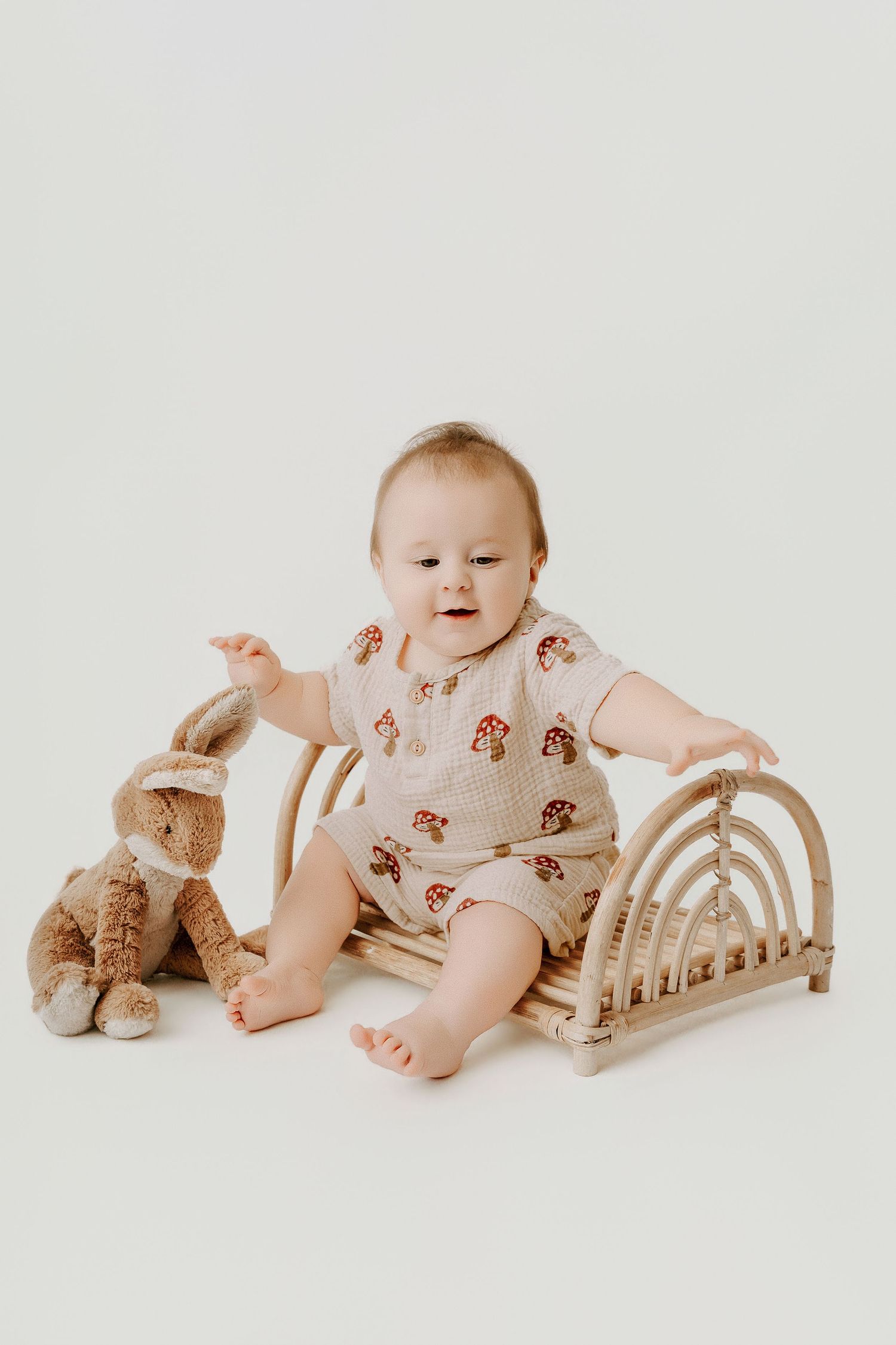 Baby in mushroom print romper sits on rattan chair with stuffed bunny toy on white background.