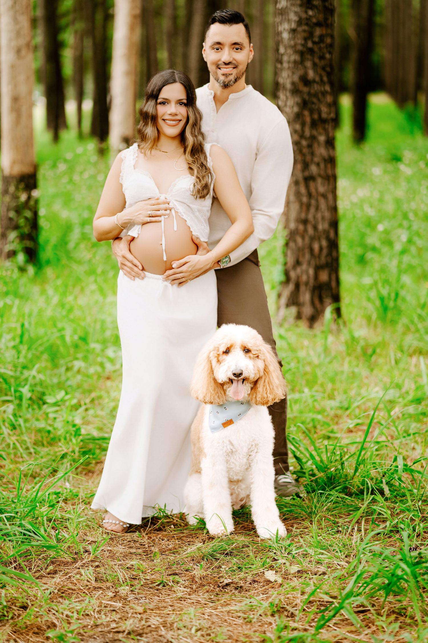 A couple in white clothing poses with their fluffy white dog in a sunlit forest setting.
