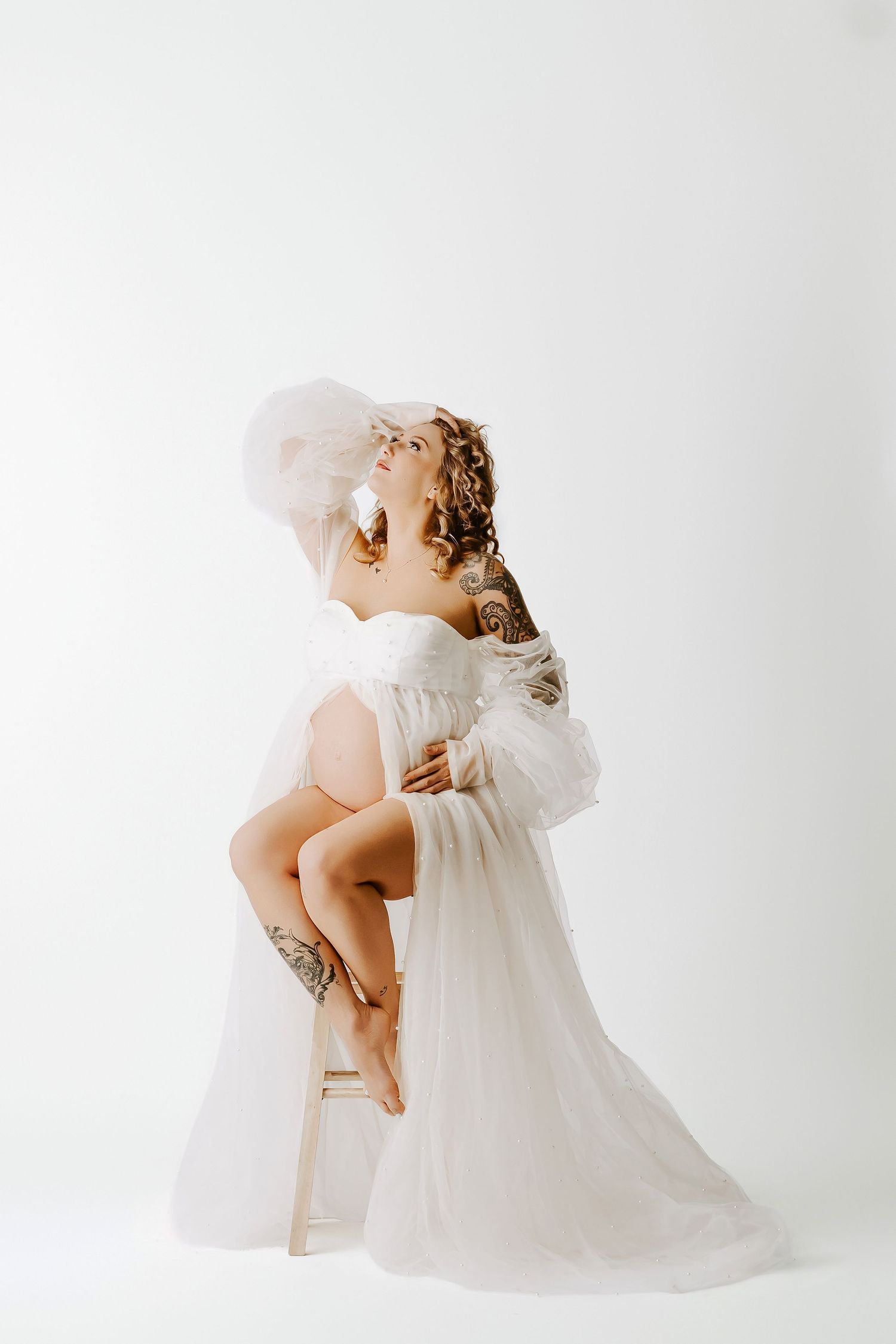 Pregnant woman in white sheer gown with tulle hat sits gracefully on stool, showcasing tattoos against white background.