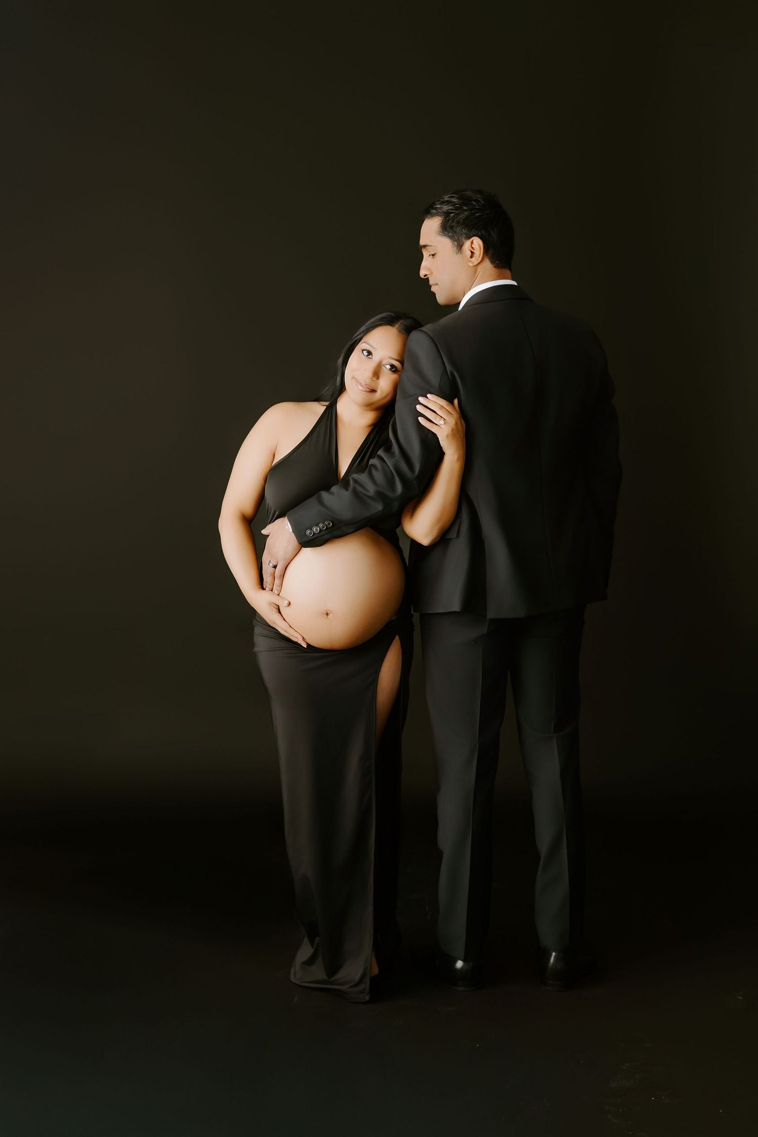 Pregnant woman in black dress poses with partner in suit for elegant maternity photo shoot on dark background.