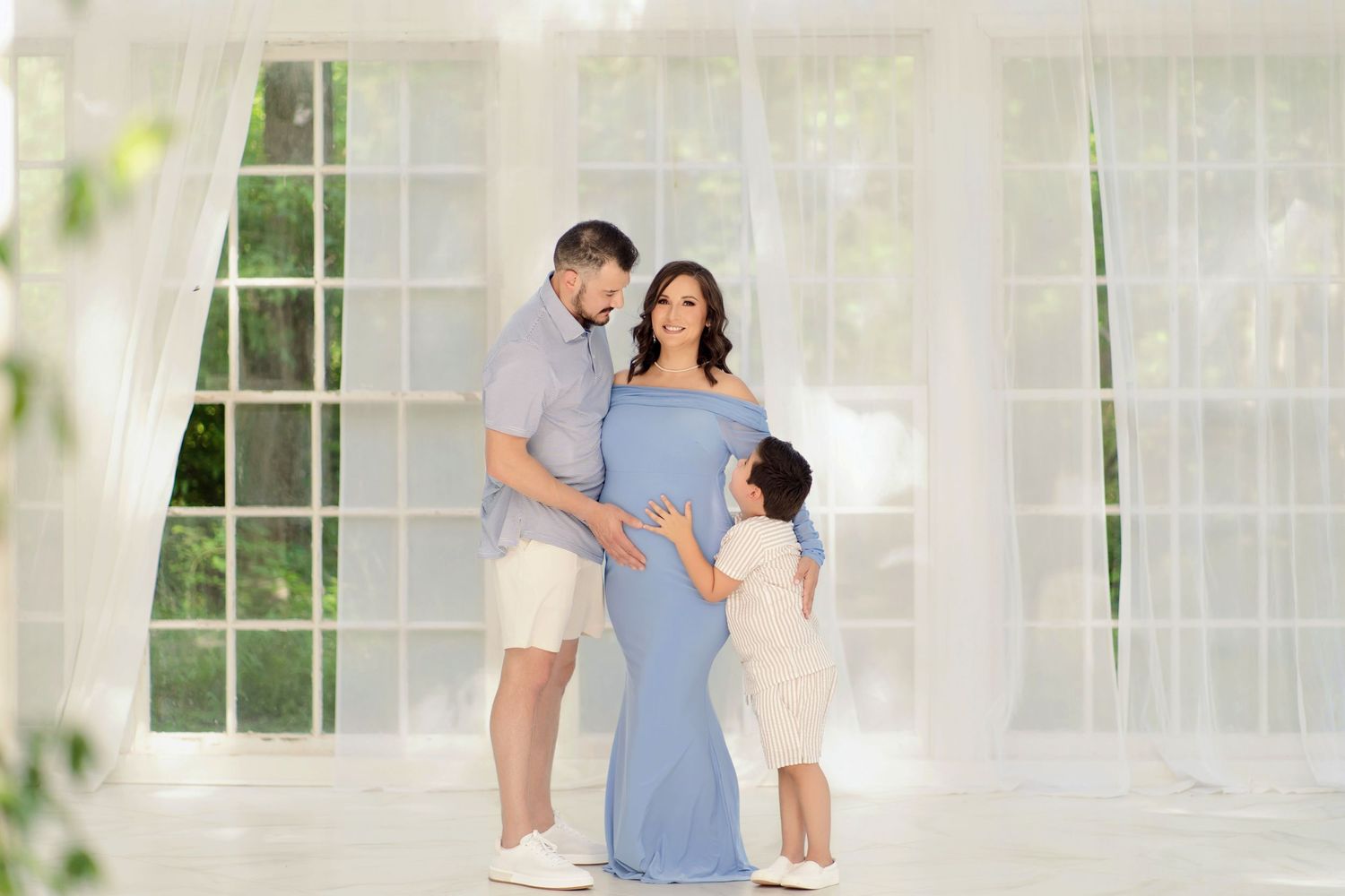 A family maternity photo shoot with a pregnant woman in a blue gown, man, and child posing in a bright white studio.