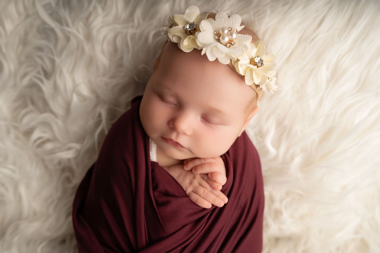 Sleeping newborn baby girl wrapped in burgundy swaddle with floral headband on white fluffy backdrop.