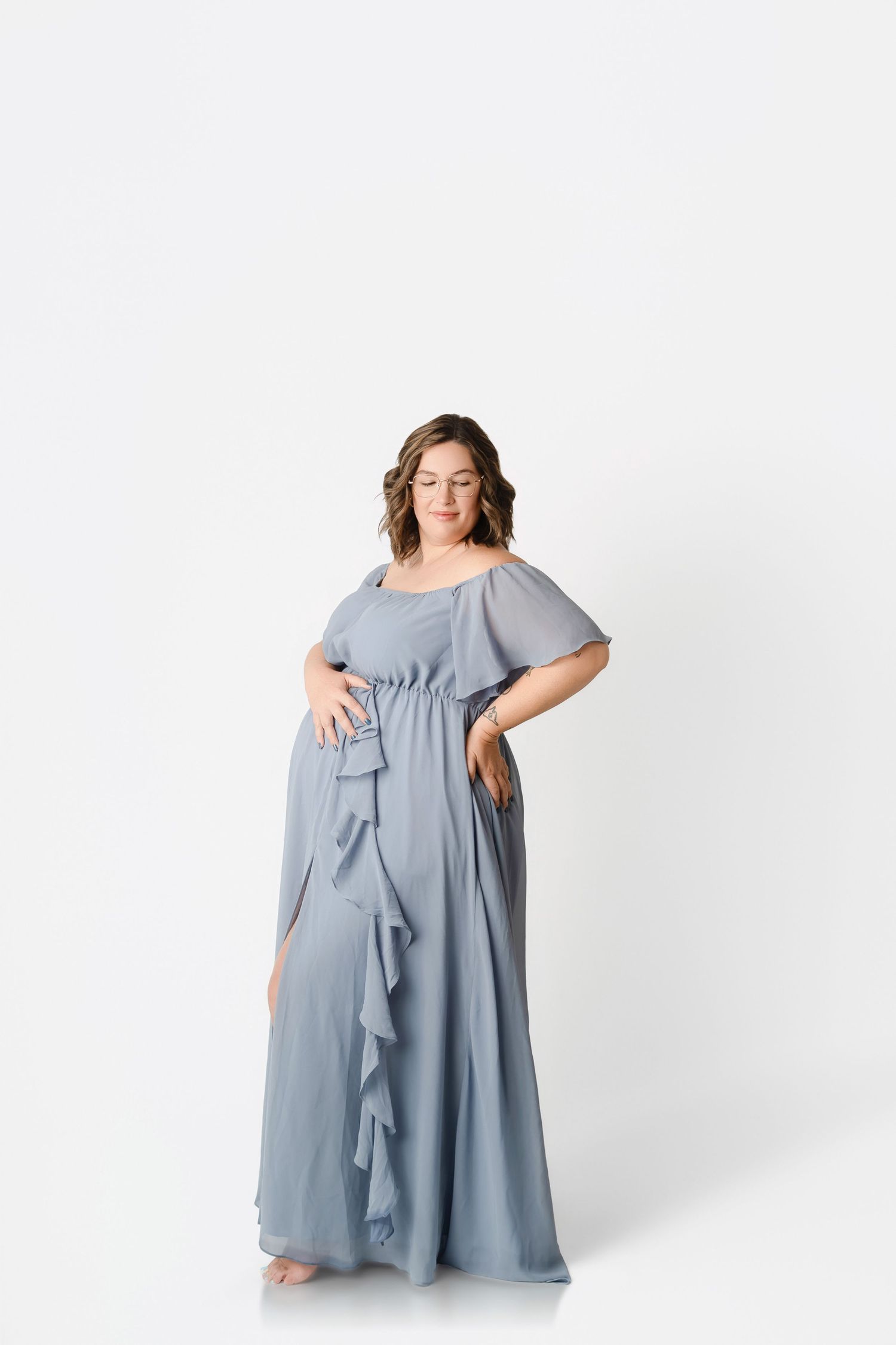 Pregnant woman in elegant dusty blue chiffon maternity gown with ruffle details, posing against white background.