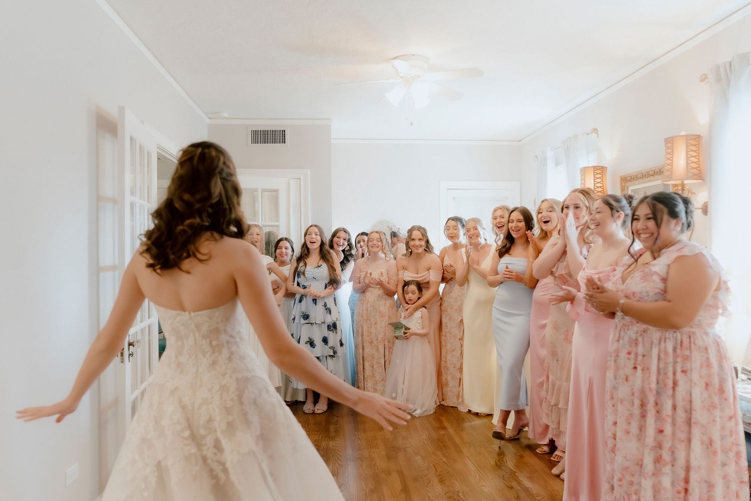 Bride in white gown reveals dress to excited bridesmaids in pastel dresses during a joyful first look moment indoors.