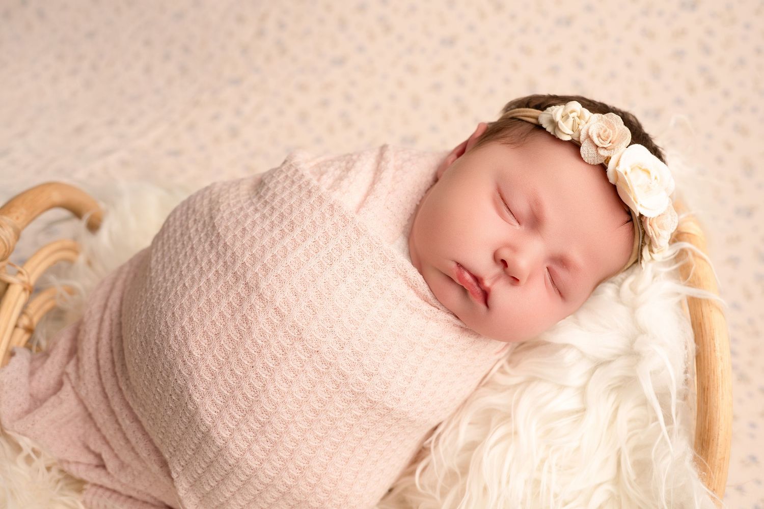 Sleeping newborn baby girl swaddled in pink wrap with floral headband in a basket.