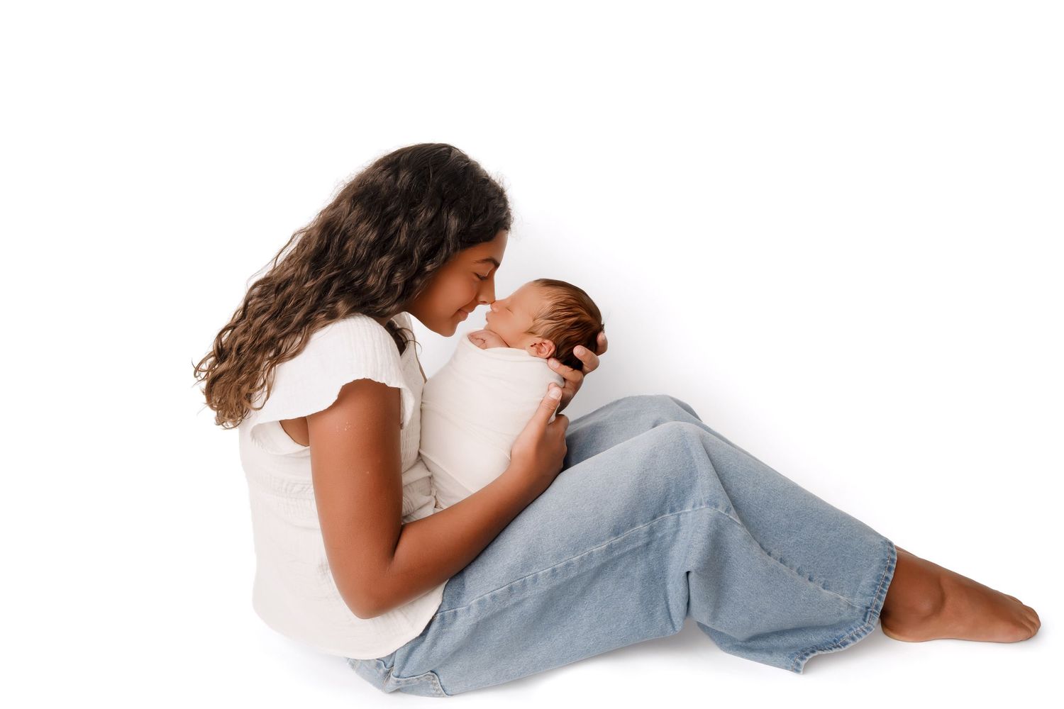 A mother sitting on white background cradling her newborn baby, wearing white top and jeans.